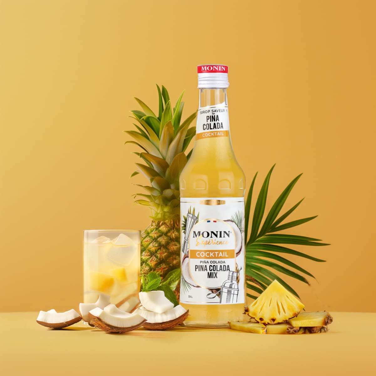 Monin Piña Colada mix with a pineapple, coconut flakes and an iced glass on yellow