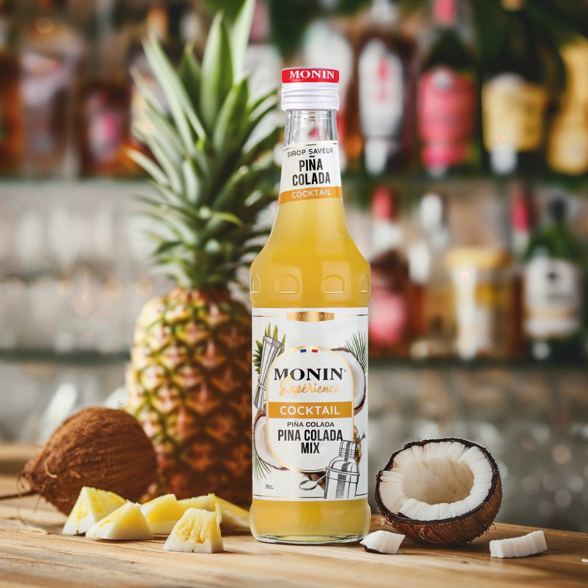 Monin Piña Colada bottle with pineapple and coconut halves on a bar with bottles behind