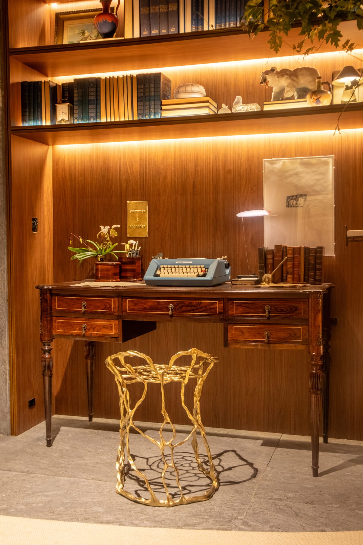 Antique writing desk with a blue typewriter and a gilded wire stool below lit shelves