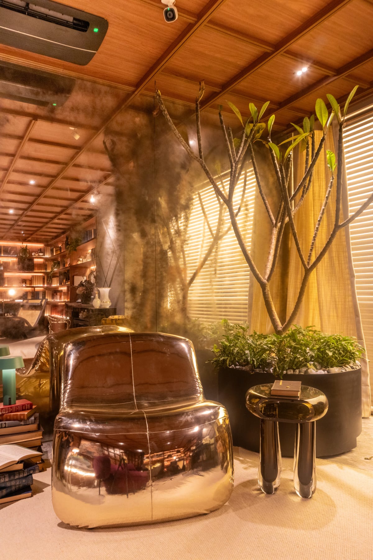 Polished bronze lounge chair and side table beside a potted tree and bookshelves