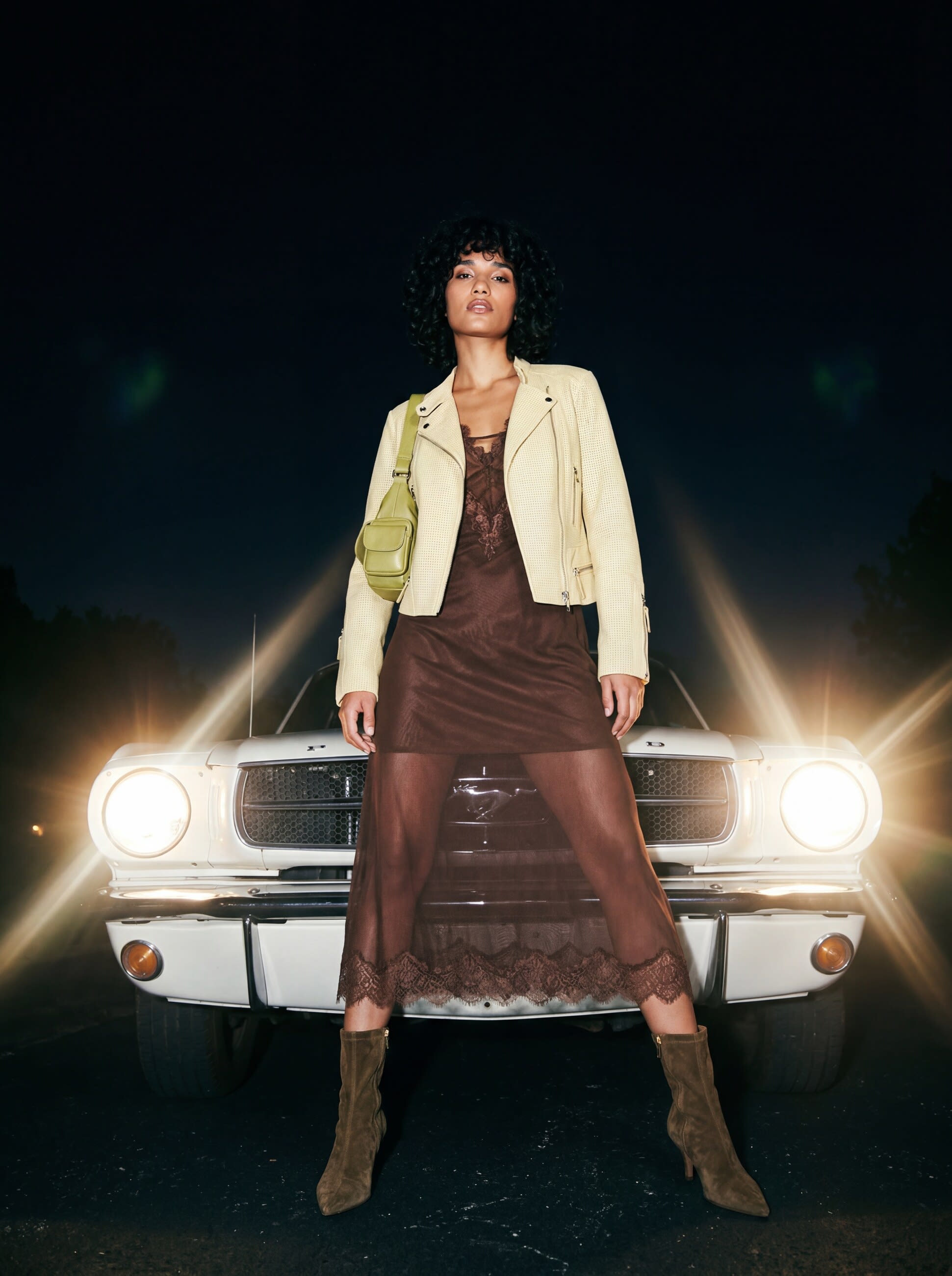 A model in a cream leather jacket, sheer brown slip dress and suede boots before Mustang headlights