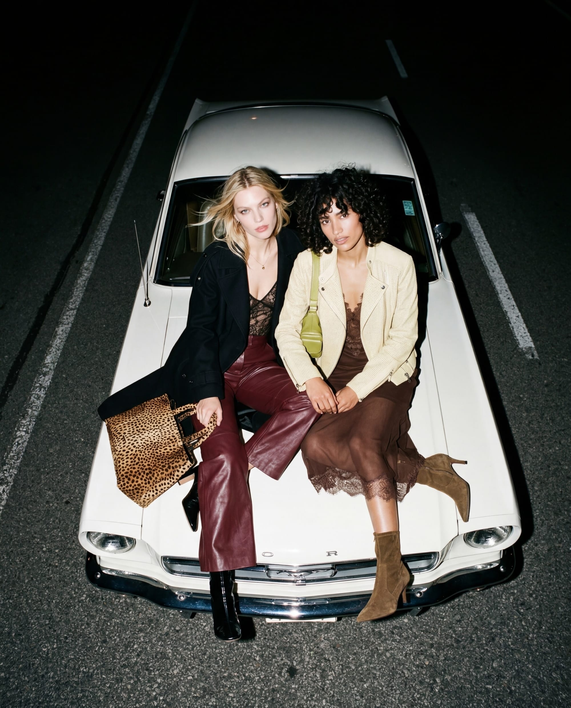 Two models with leopard and green bags sitting on a white Mustang hood at night