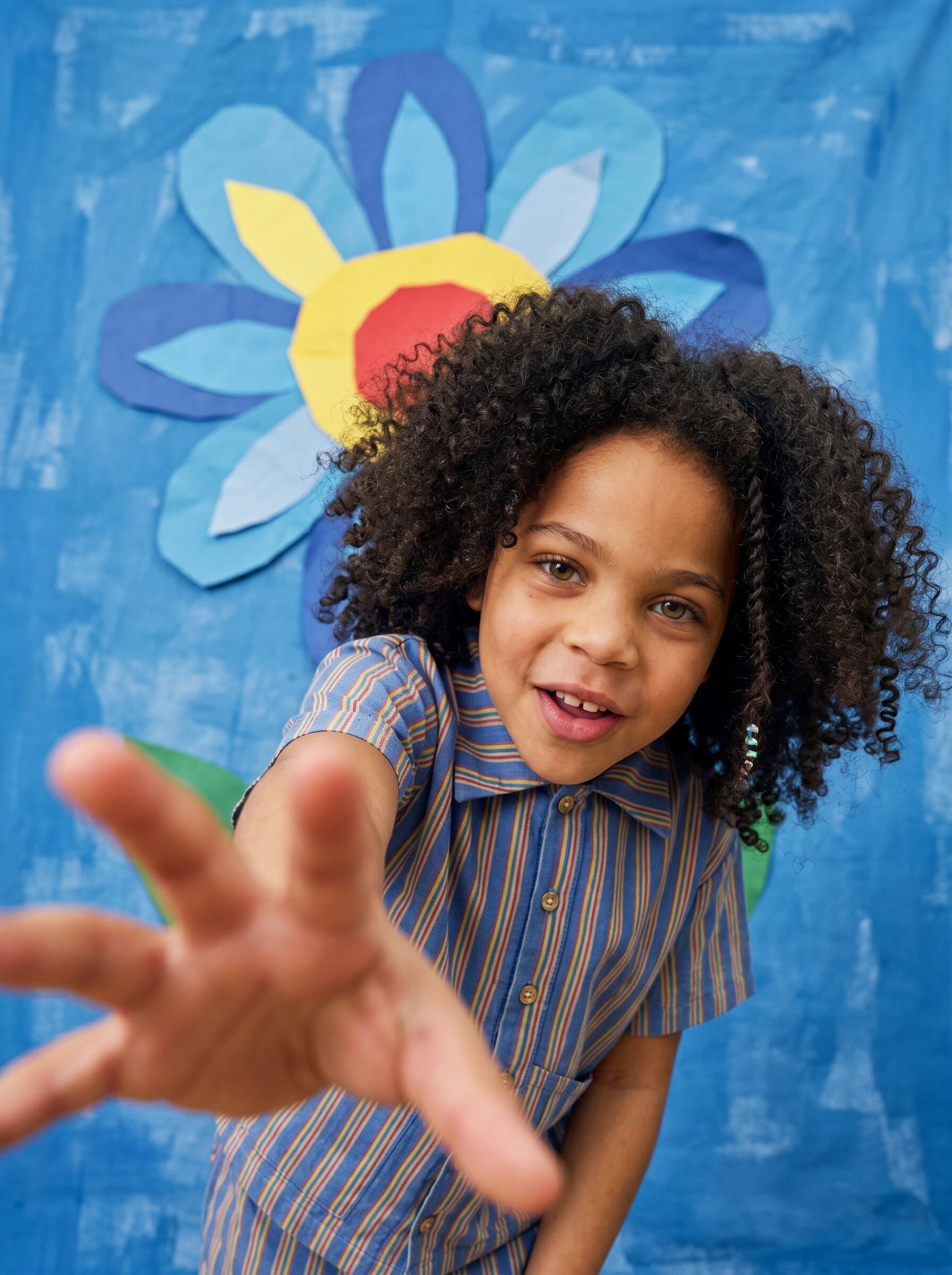 A child in a striped short sleeve shirt reaching toward the lens before a cut paper flower