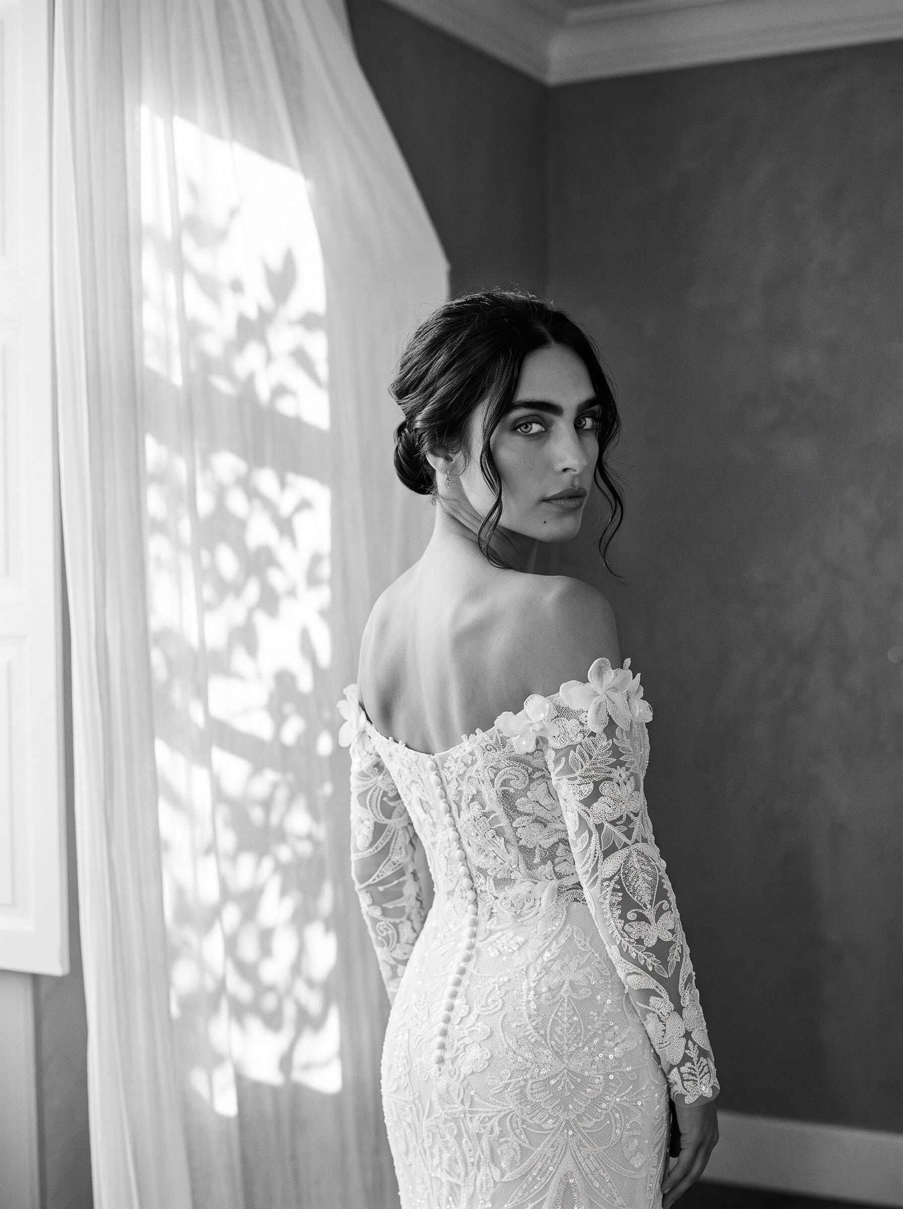 Black and white back view of a buttoned lace bridal gown by a sheer curtained window