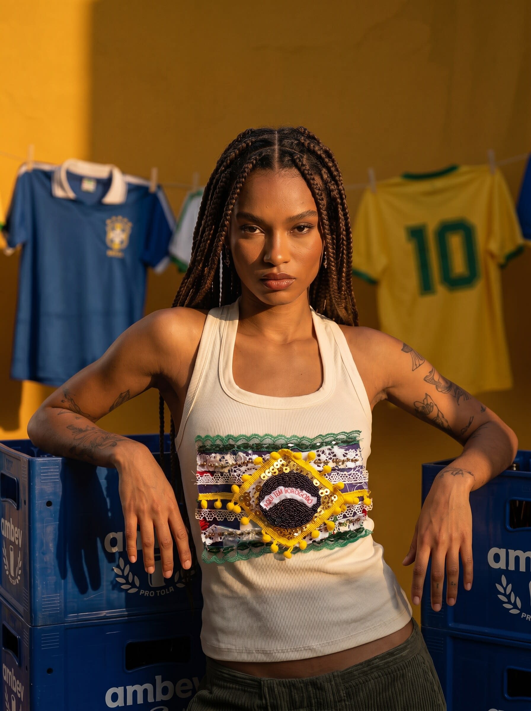 A model in a beaded Brazil flag tank top leaning on blue crates beside hanging vintage jerseys