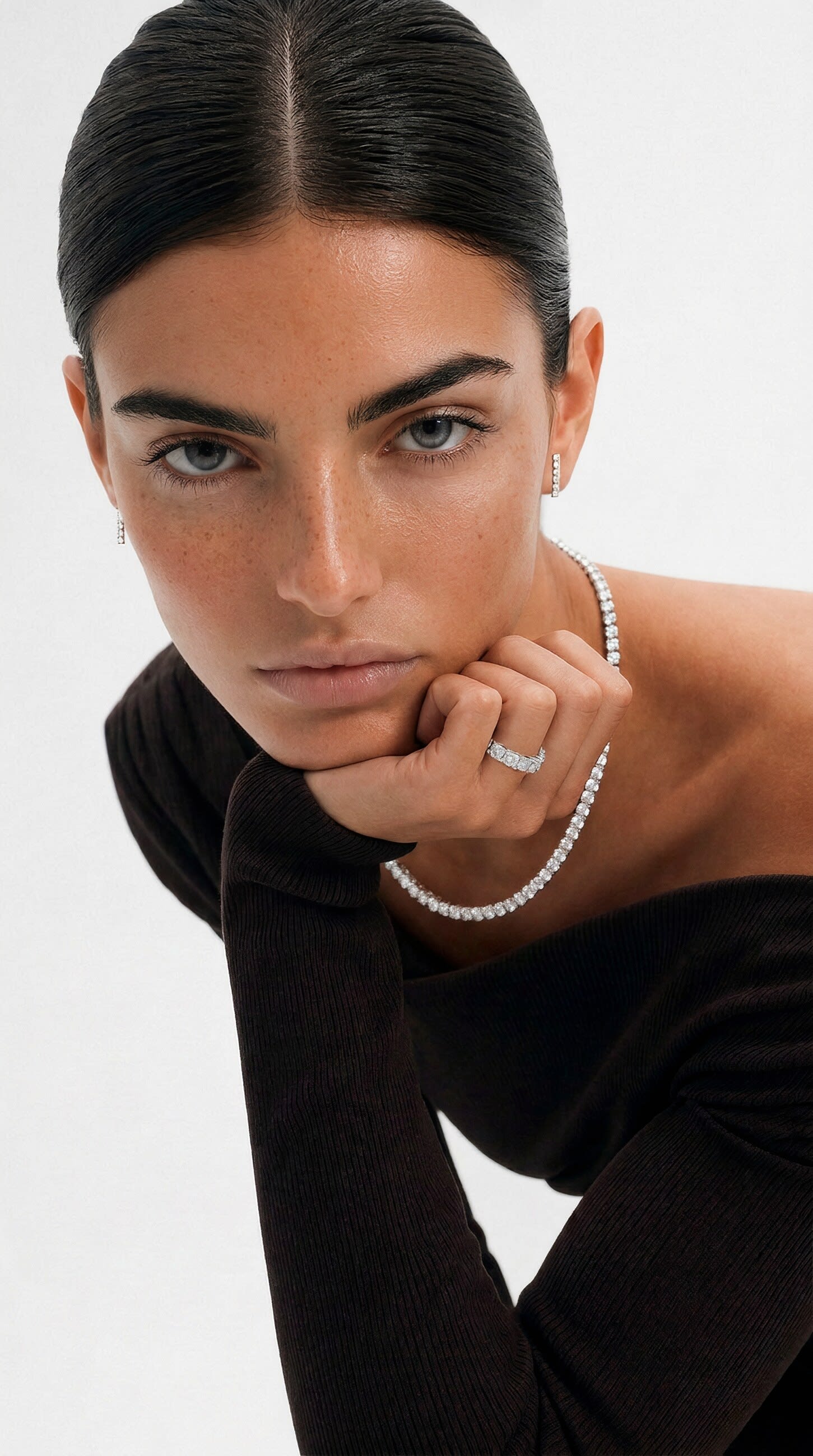 A model in a black knit resting on folded hands, wearing a diamond tennis necklace and stacked rings