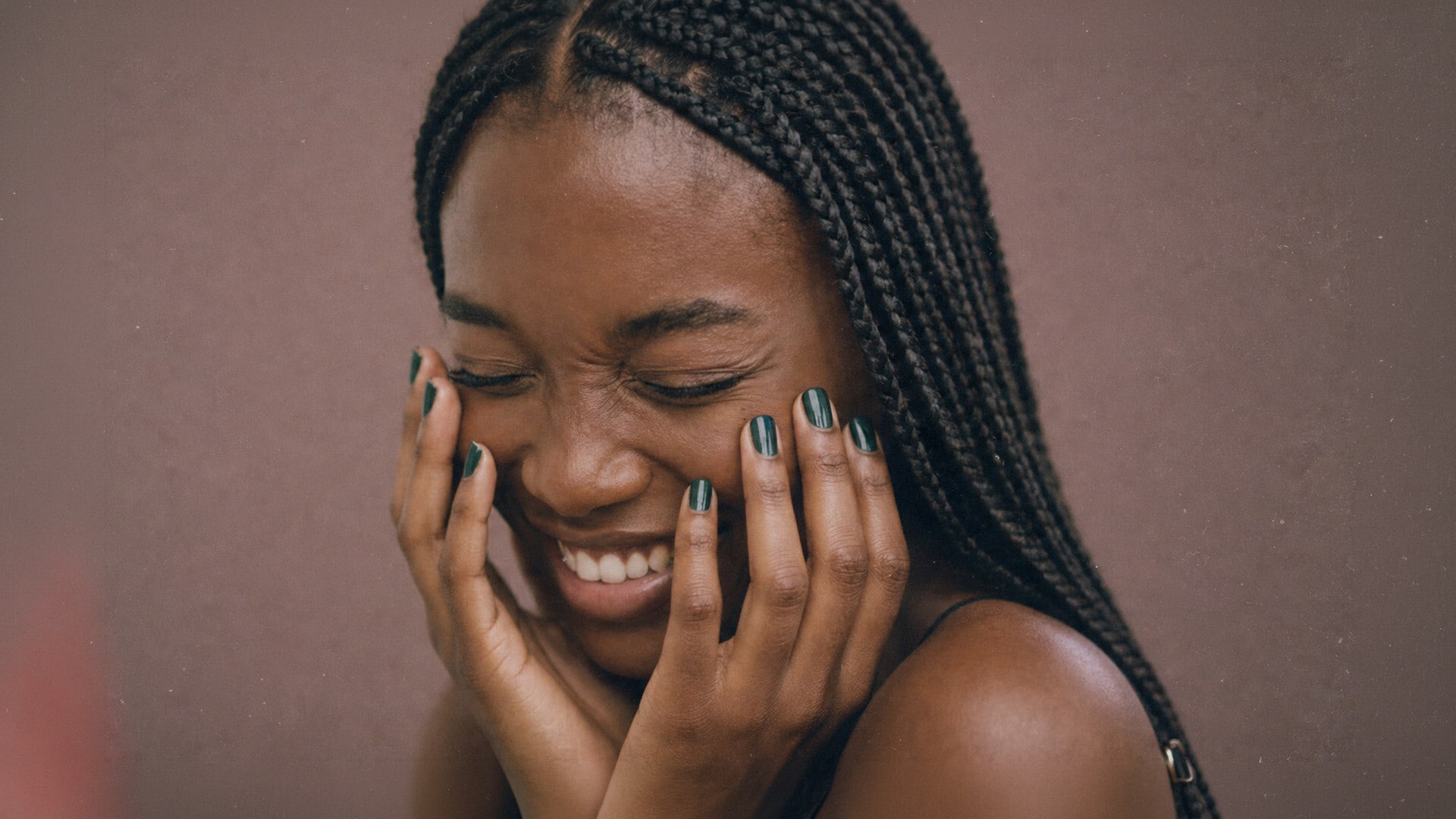 Hands with dark green nail polish held to the cheeks in a laugh on a mauve backdrop