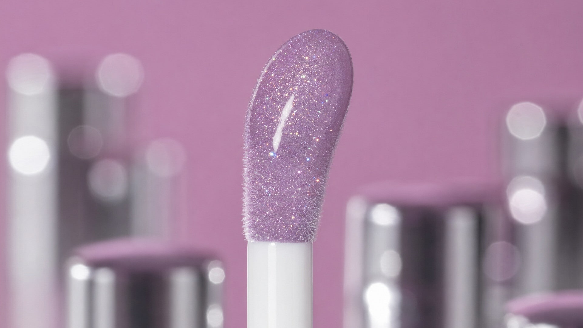 Glitter lilac lip gloss wand standing in front of blurred silver cosmetic caps on pink