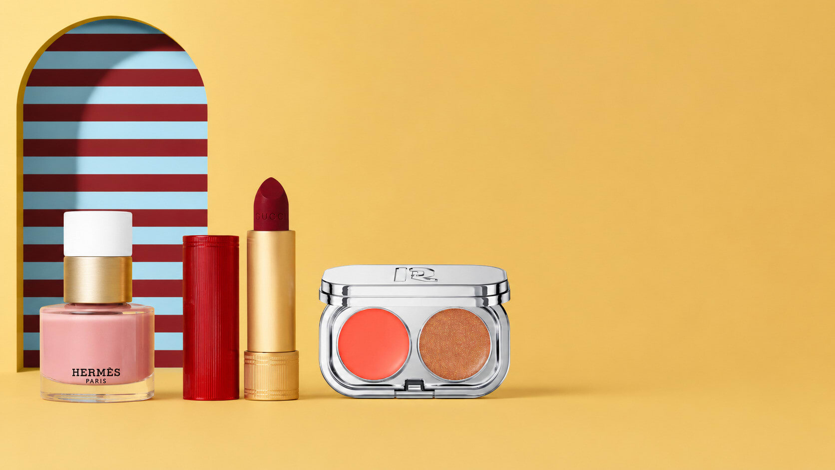 Pink nail polish, a red lipstick and a blush duo before a striped arch on yellow
