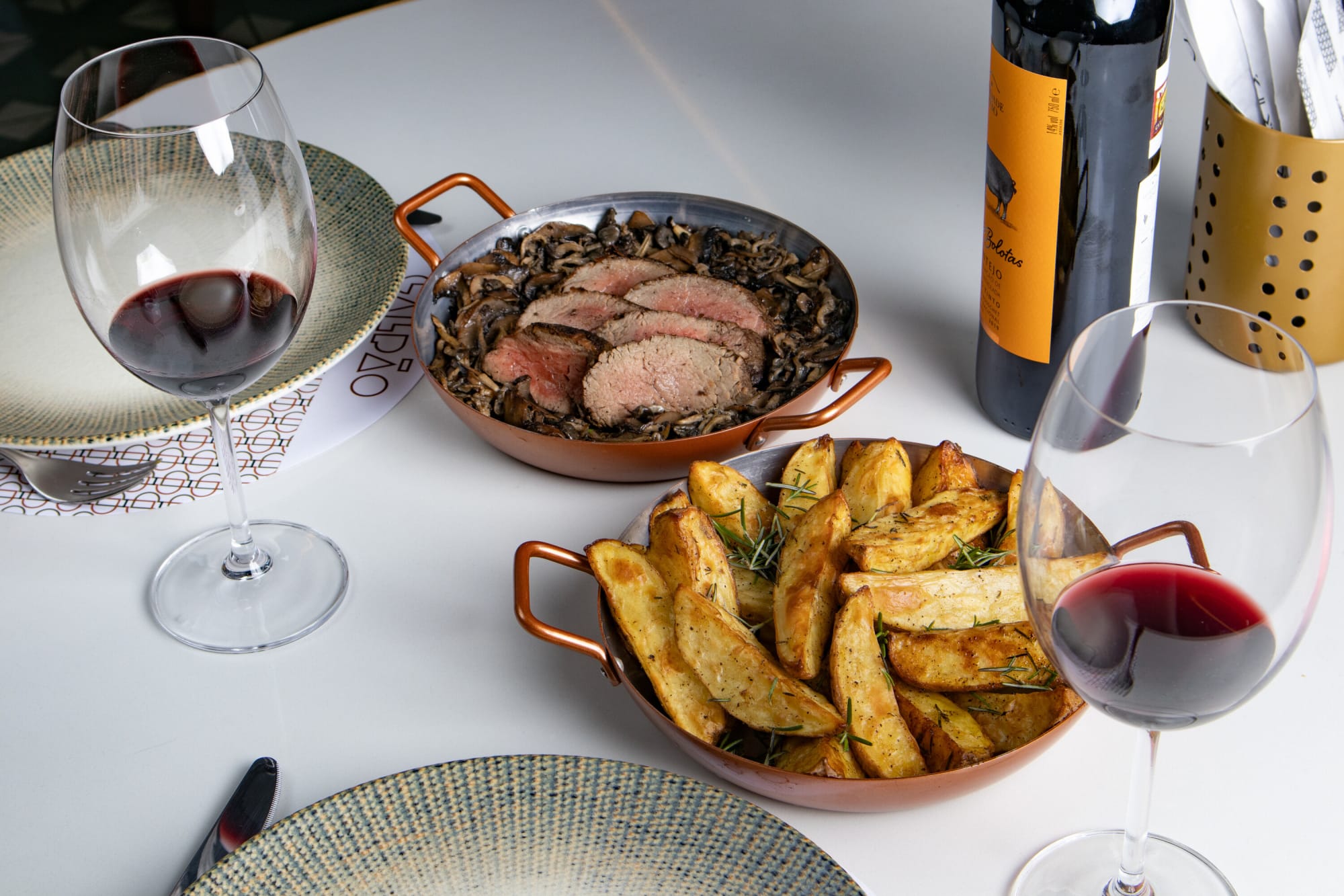 Sliced steak on greens and potato wedges in copper pans with red wine on marble