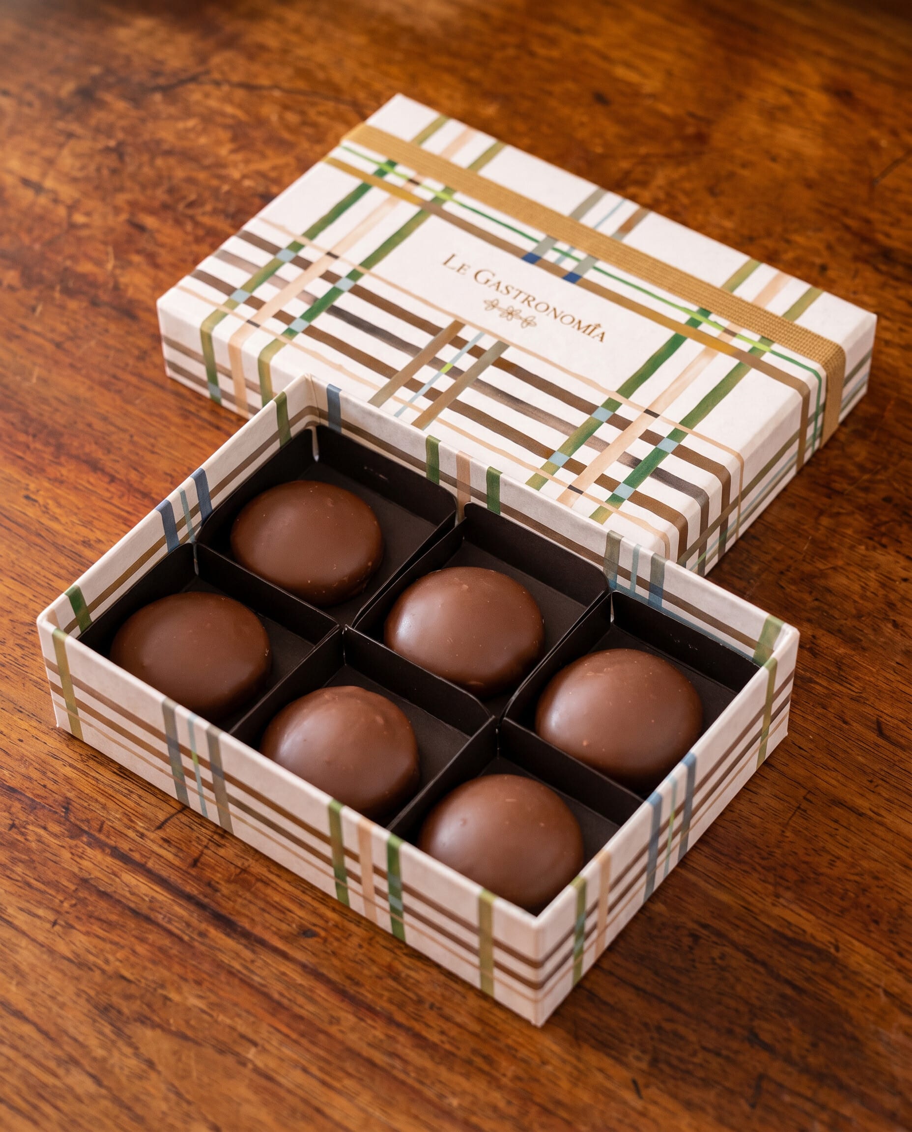 Open plaid Le Gastronomia box of six chocolate bonbons on a dark wood table
