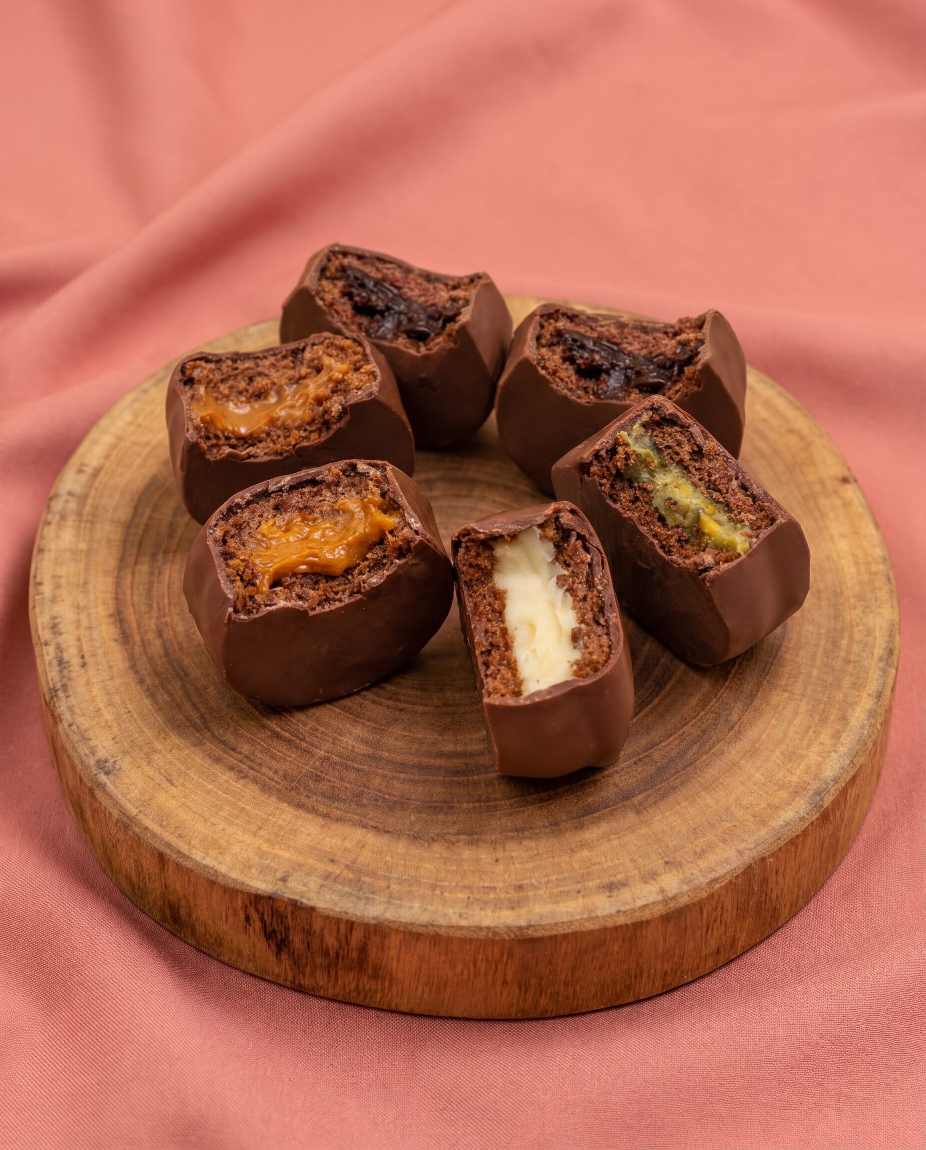 Halved chocolate bonbons with caramel, pistachio and cream fillings on a wood board over pink linen