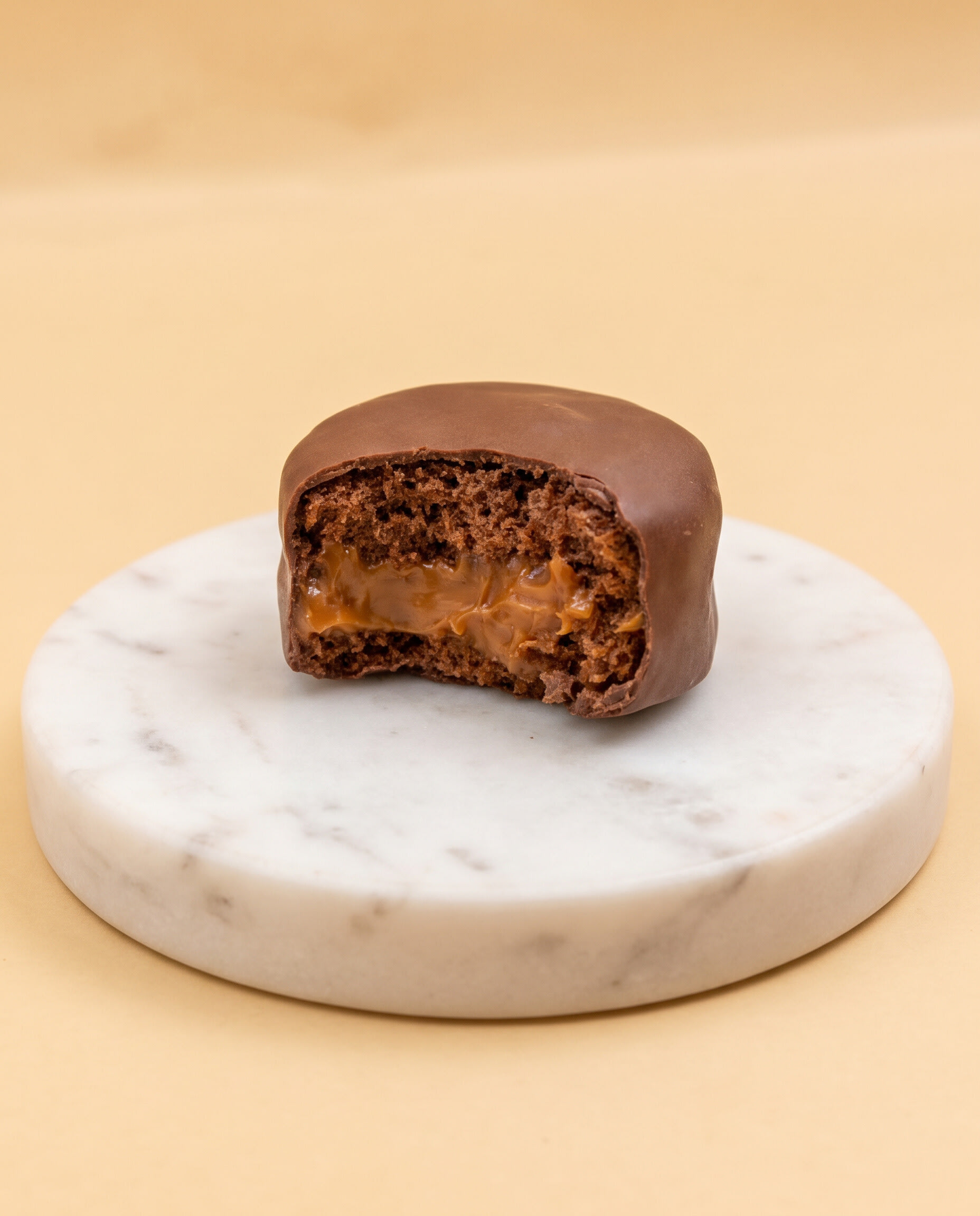 Single bitten chocolate bonbon with caramel centre on a white marble disc against peach