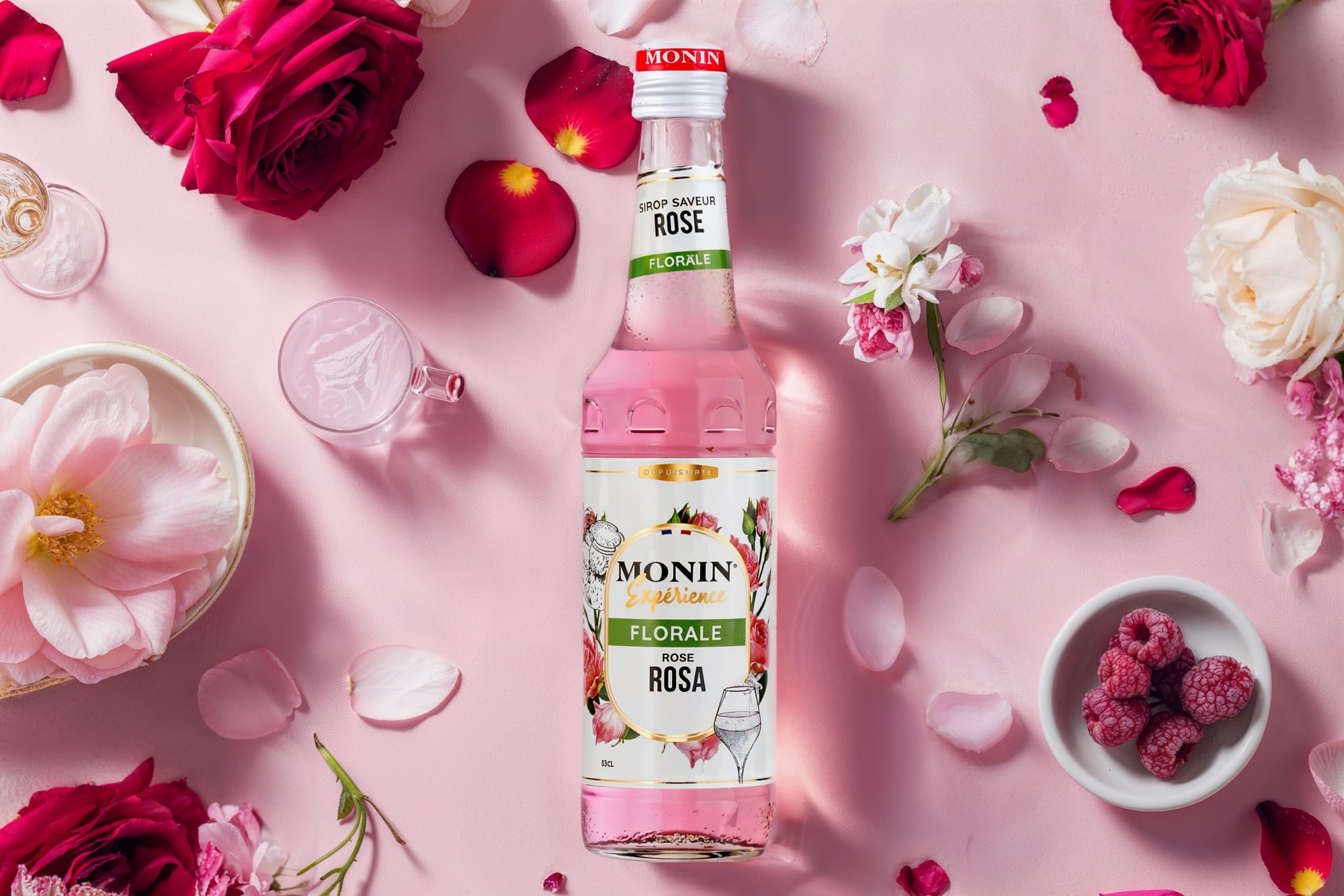 Monin rose syrup bottle with red roses, petals and raspberries on pink plaster