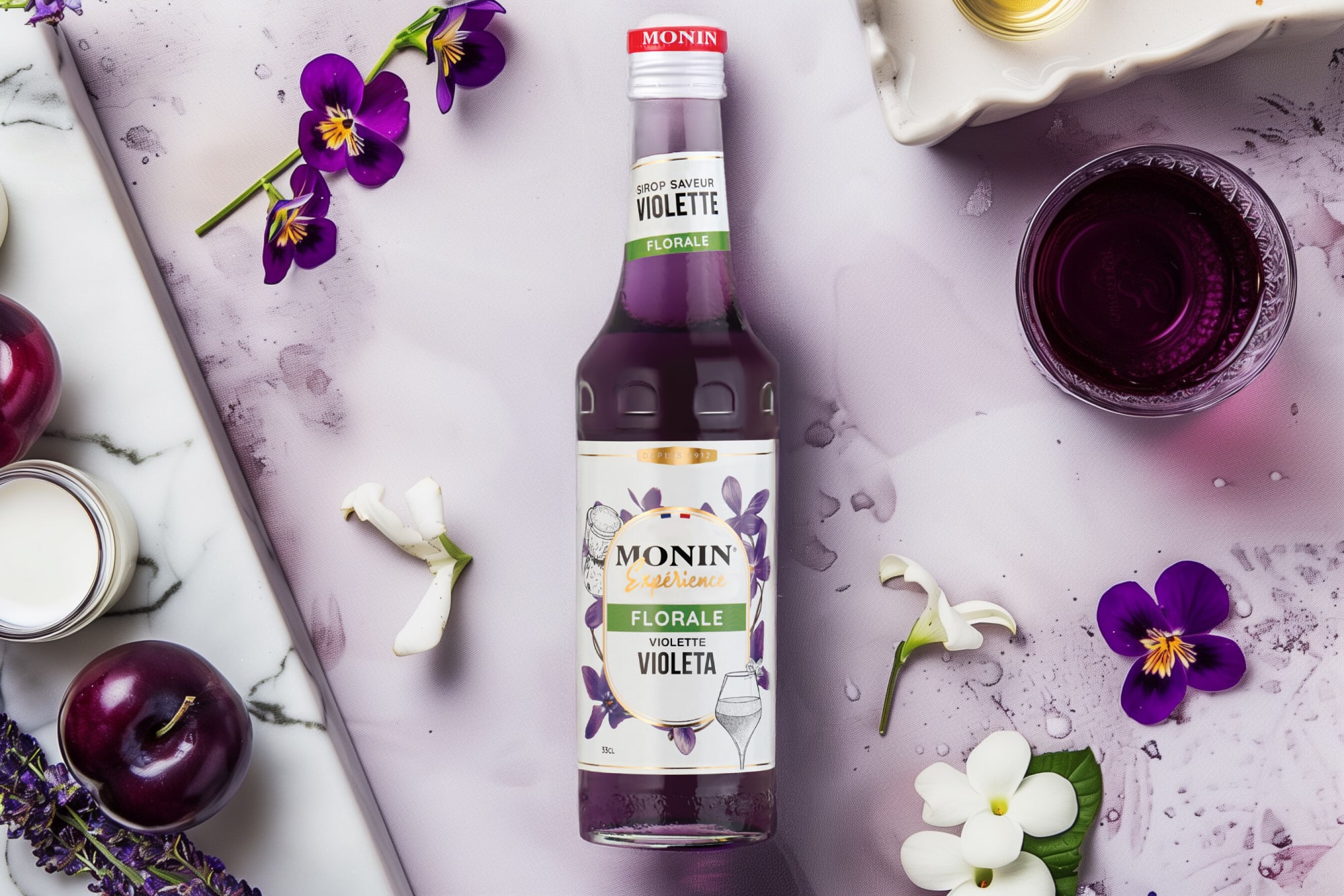 Monin violet syrup bottle with violet flowers, plums and a glass on white marble