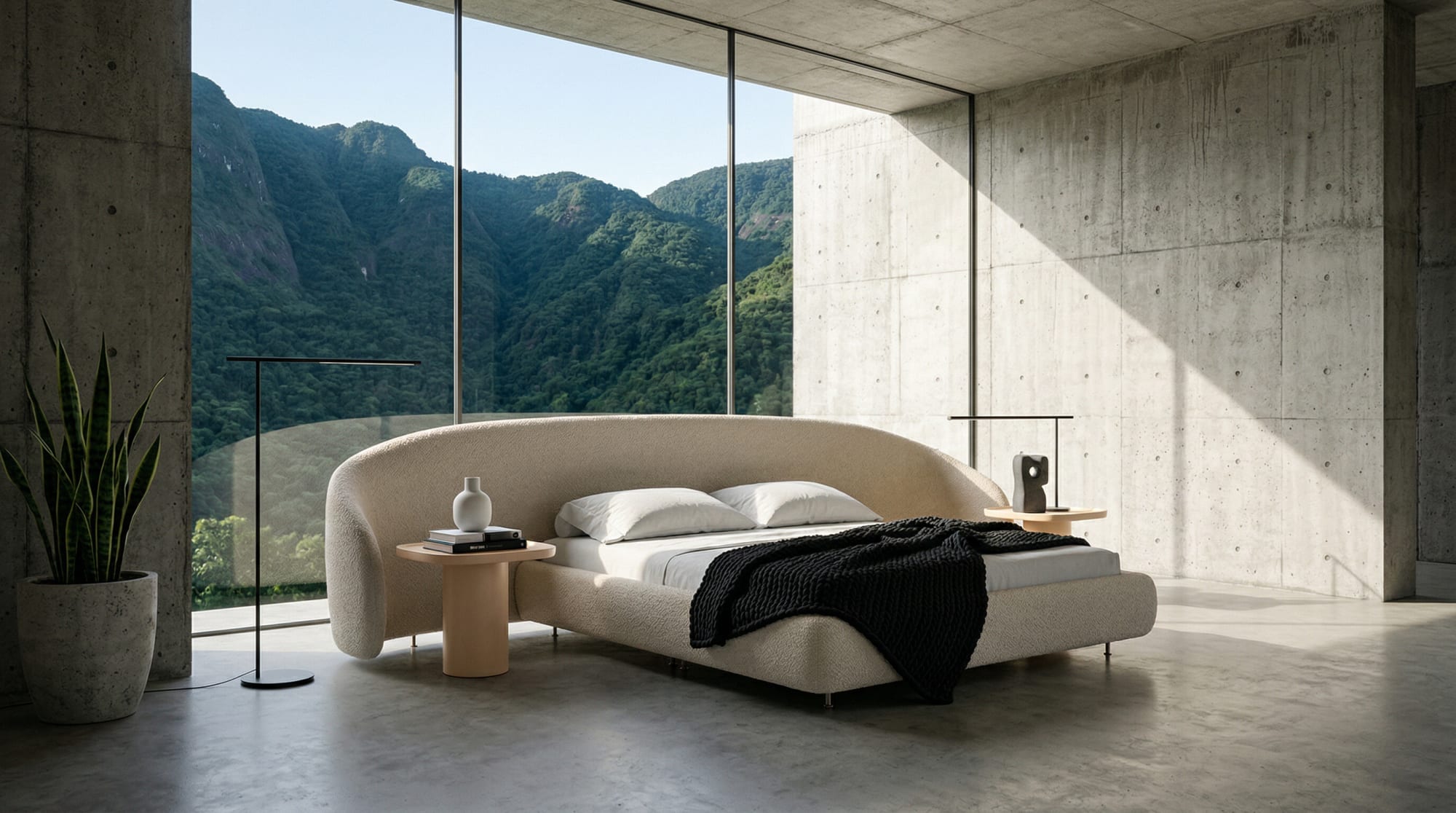 The curved Breton Malu bed with a black knit throw in a concrete room facing a green mountain