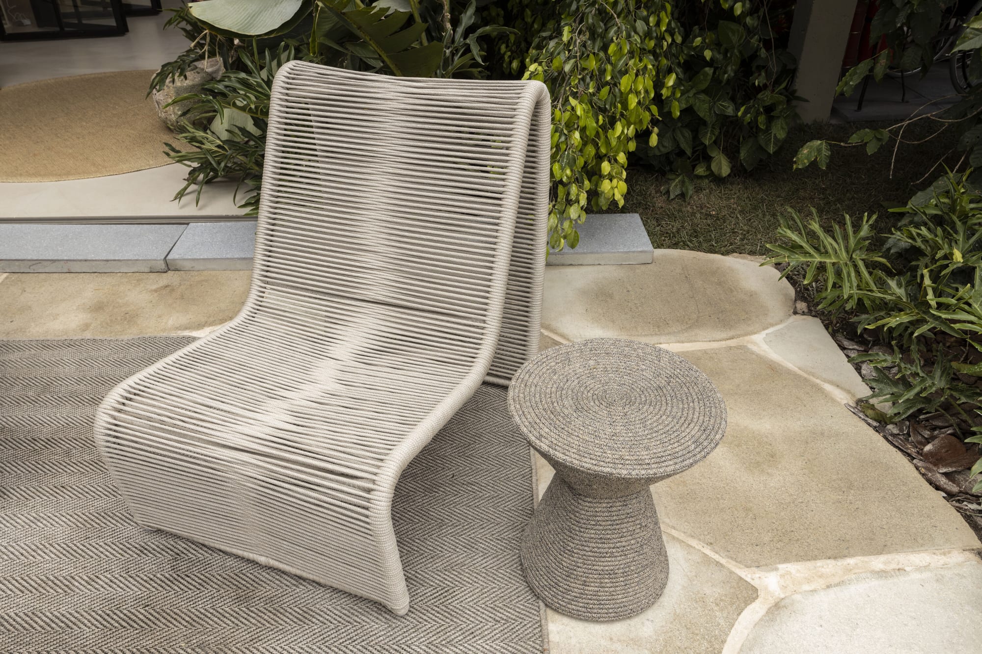 Sillón de cuerda tejida y mesa auxiliar de cuerda sobre adoquines de piedra junto a plantas tropicales