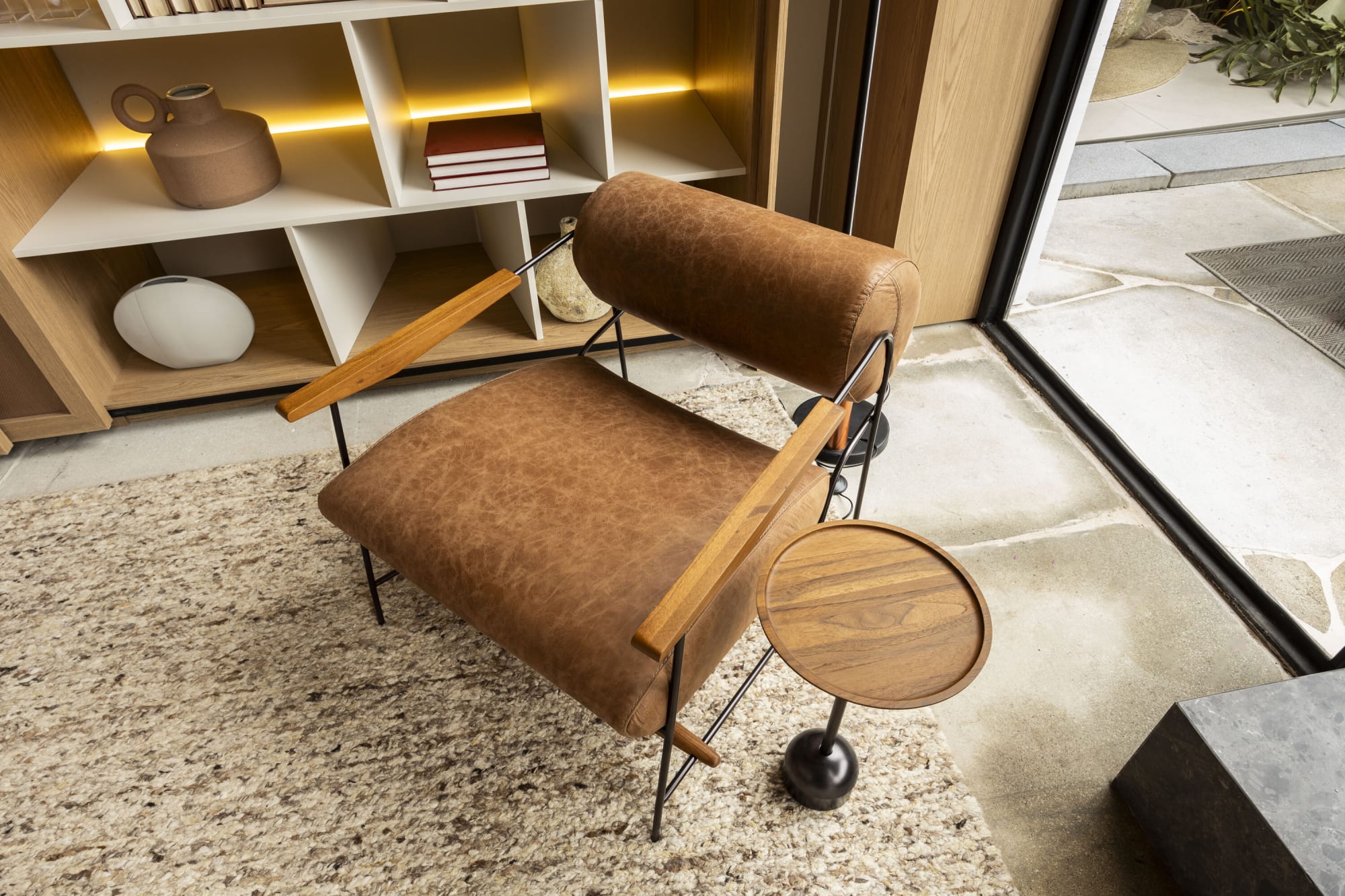 Sillón de piel camel con brazo de cojín cilíndrico y charola lateral de madera sobre piso de terrazo