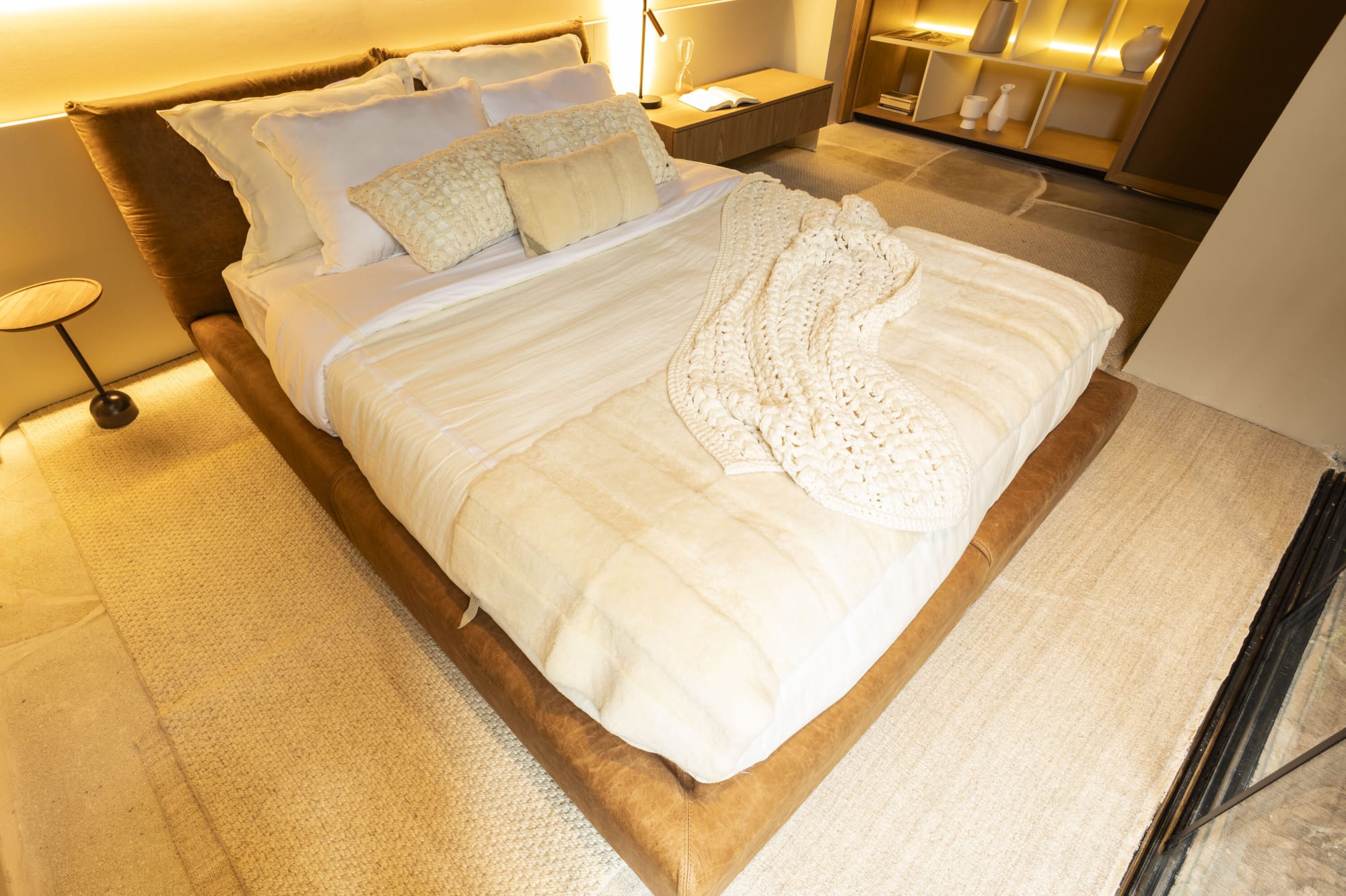 Cama blanca acolchada con manta tejida sobre una base baja de madera junto a una lámpara de piso