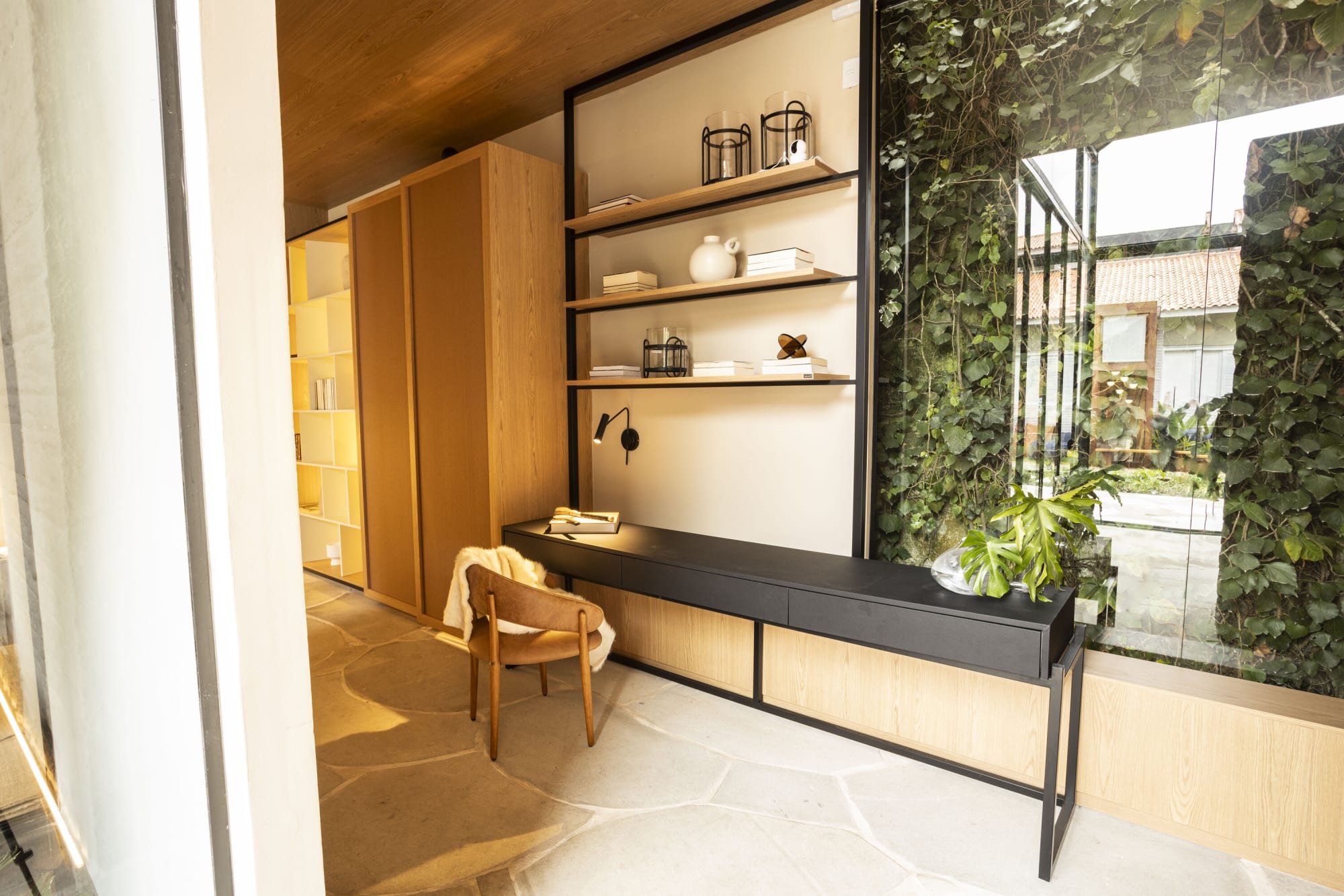 Escritorio y repisas de acero negro con una silla tejida junto a un muro de vidrio con plantas