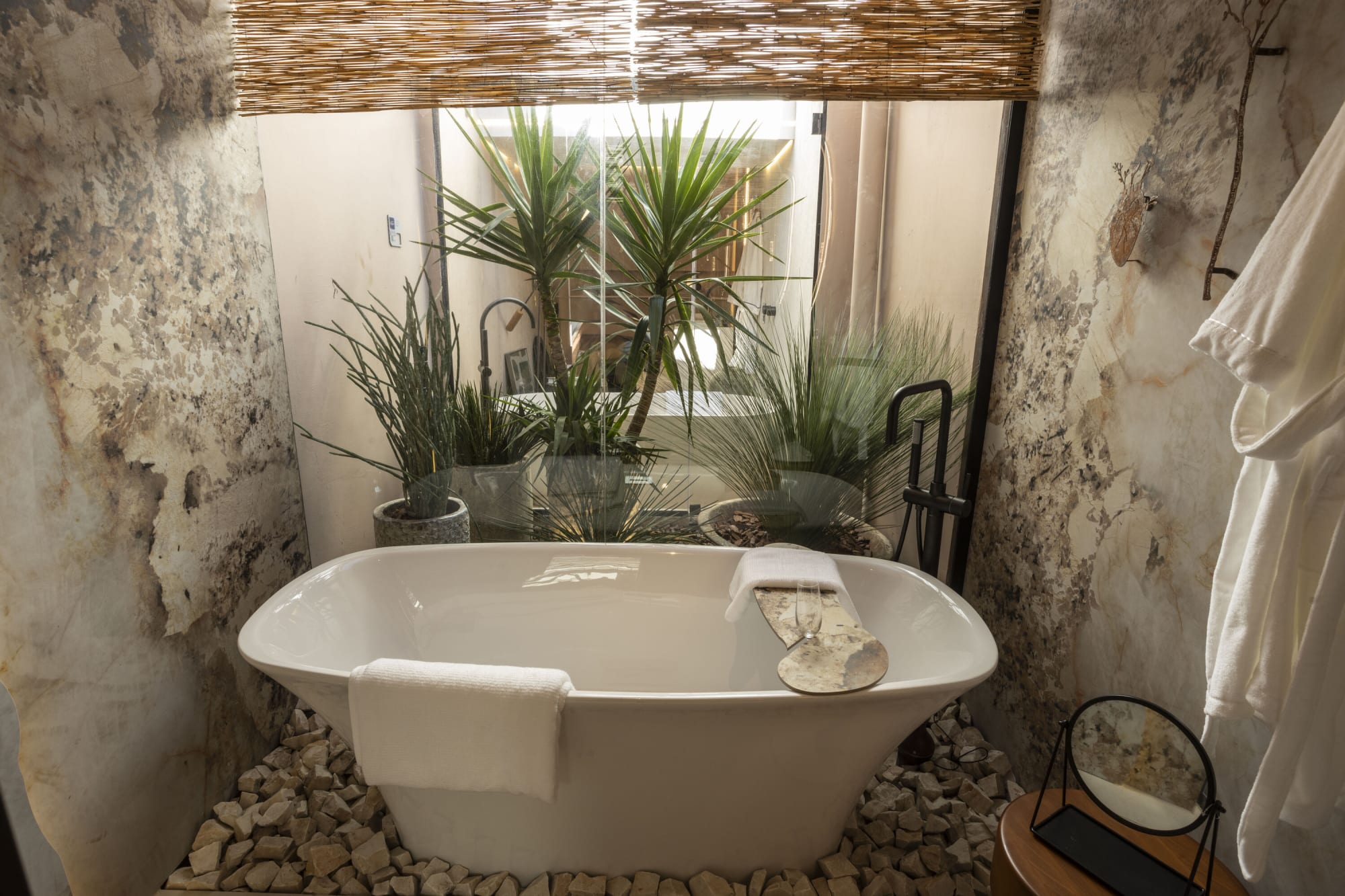 Freestanding white bathtub on a pebble bed under a planted window at CASACOR Curitiba