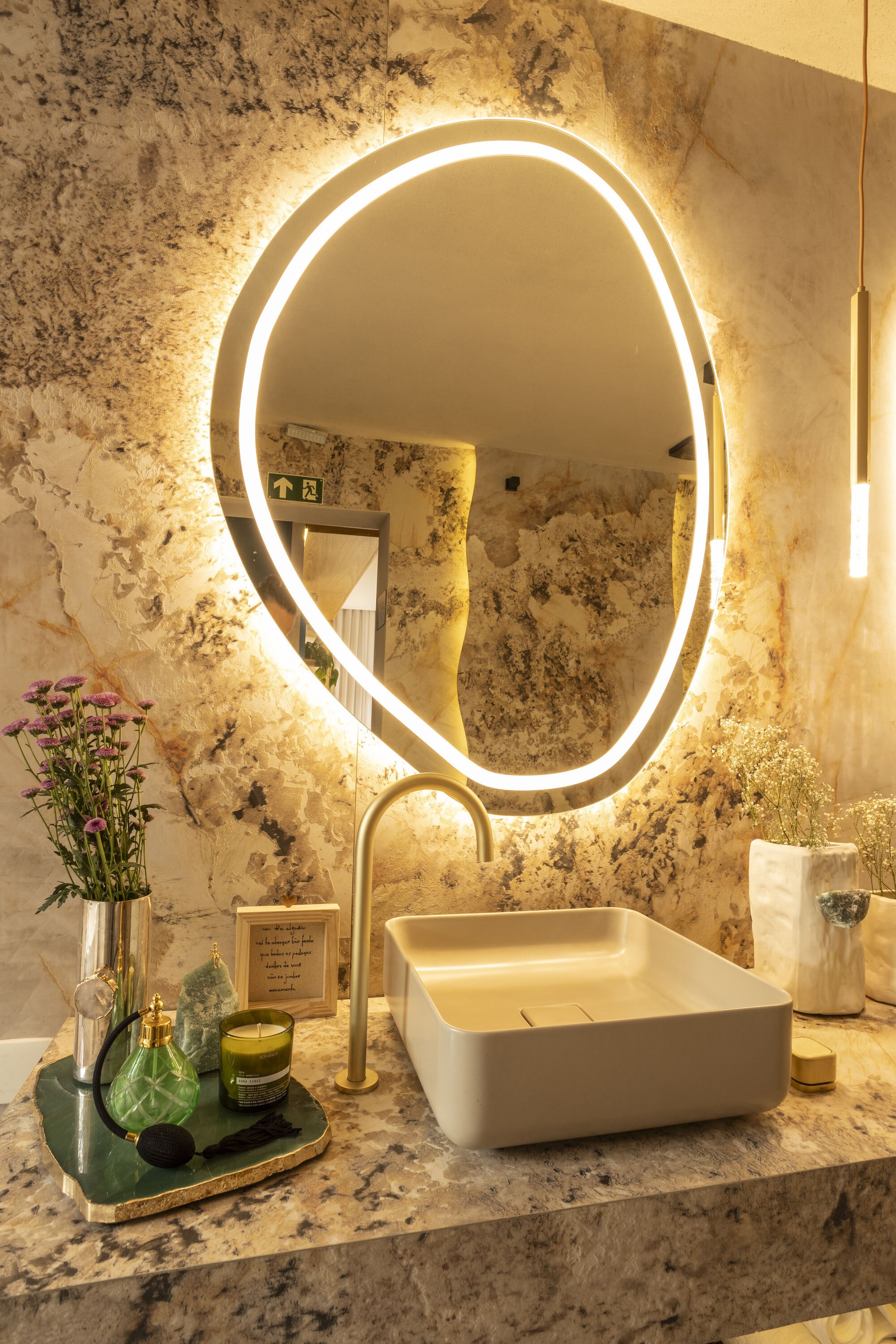 Backlit pebble shaped mirror over a white vessel basin and brass tap on granite