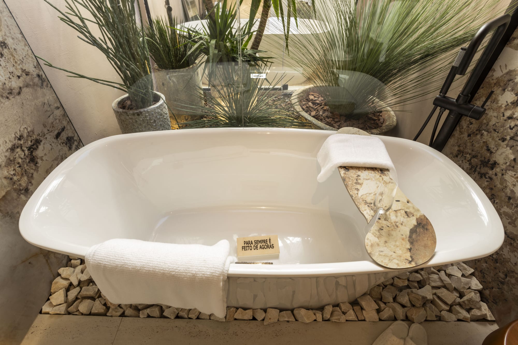 White soaking tub with a stone bath tray set in pebbles among potted grasses