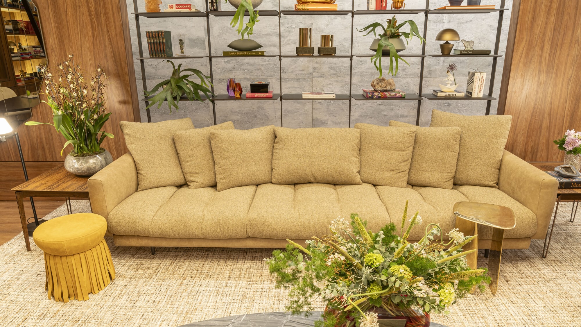 Beige sofa with mustard fringed pouf and flower arrangement against open steel shelving