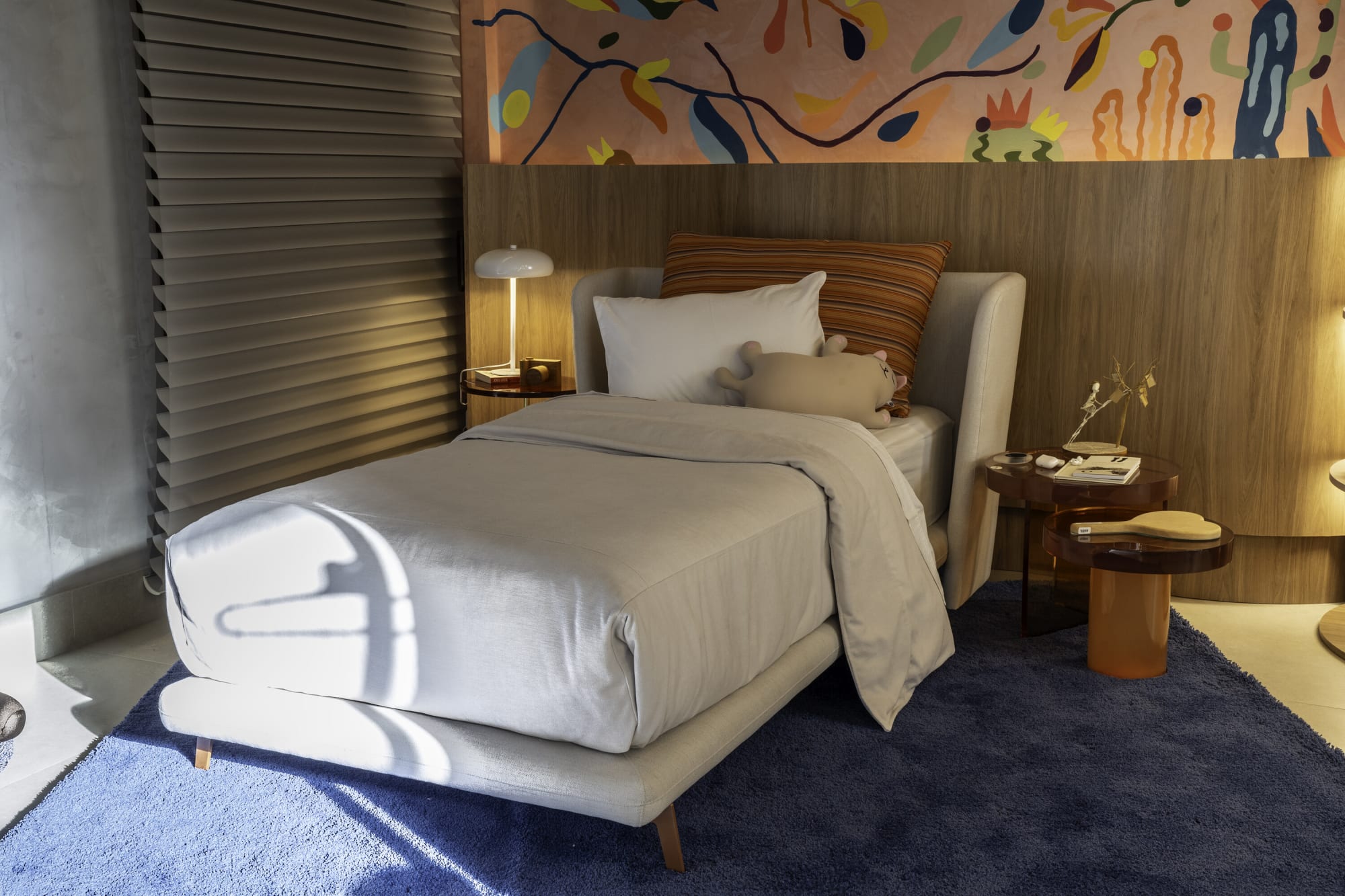 Single bed with white linen, rust cushions and a navy rug under a colourful mural frieze