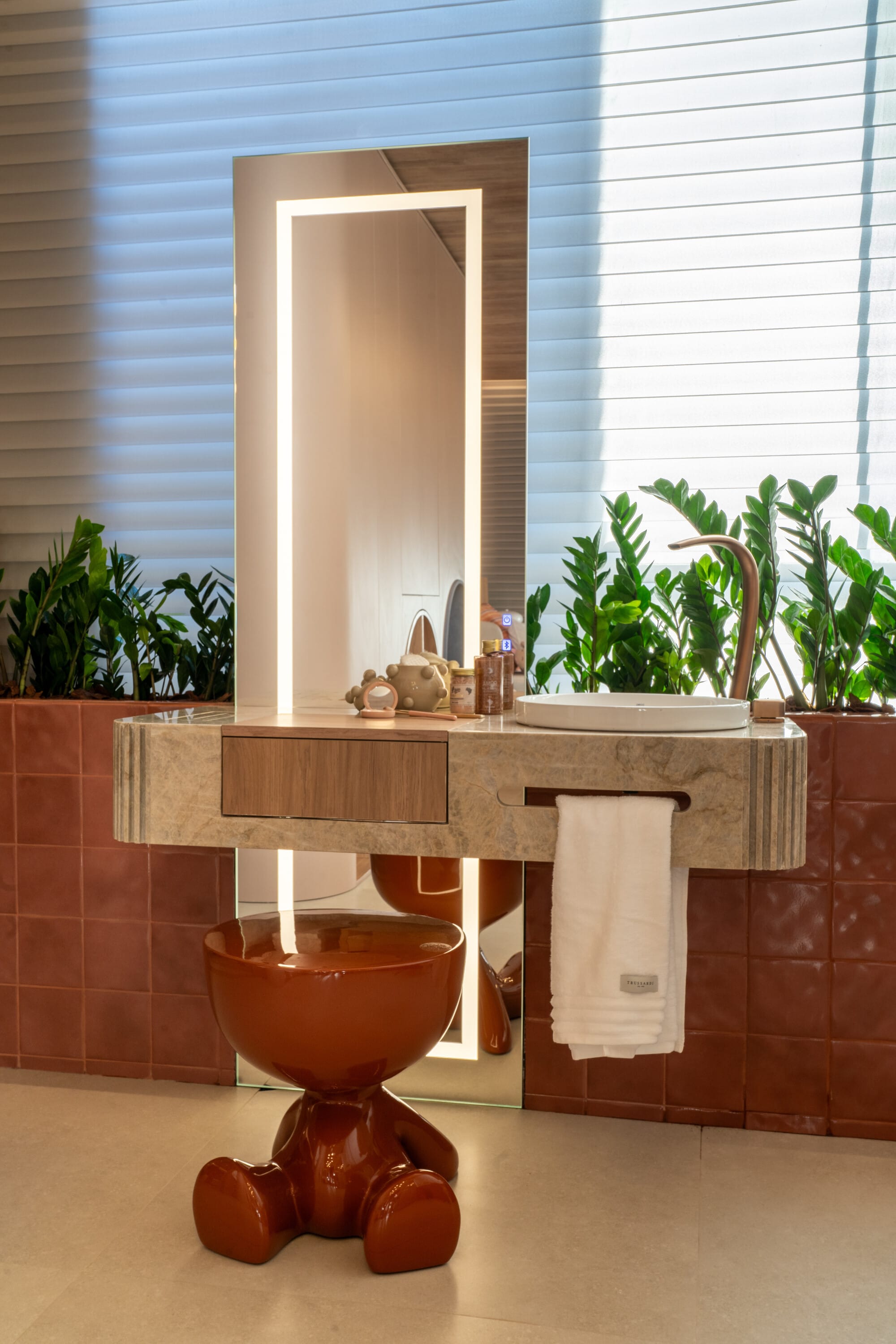 Backlit tall mirror over a stone vanity with a rust teddy bear stool