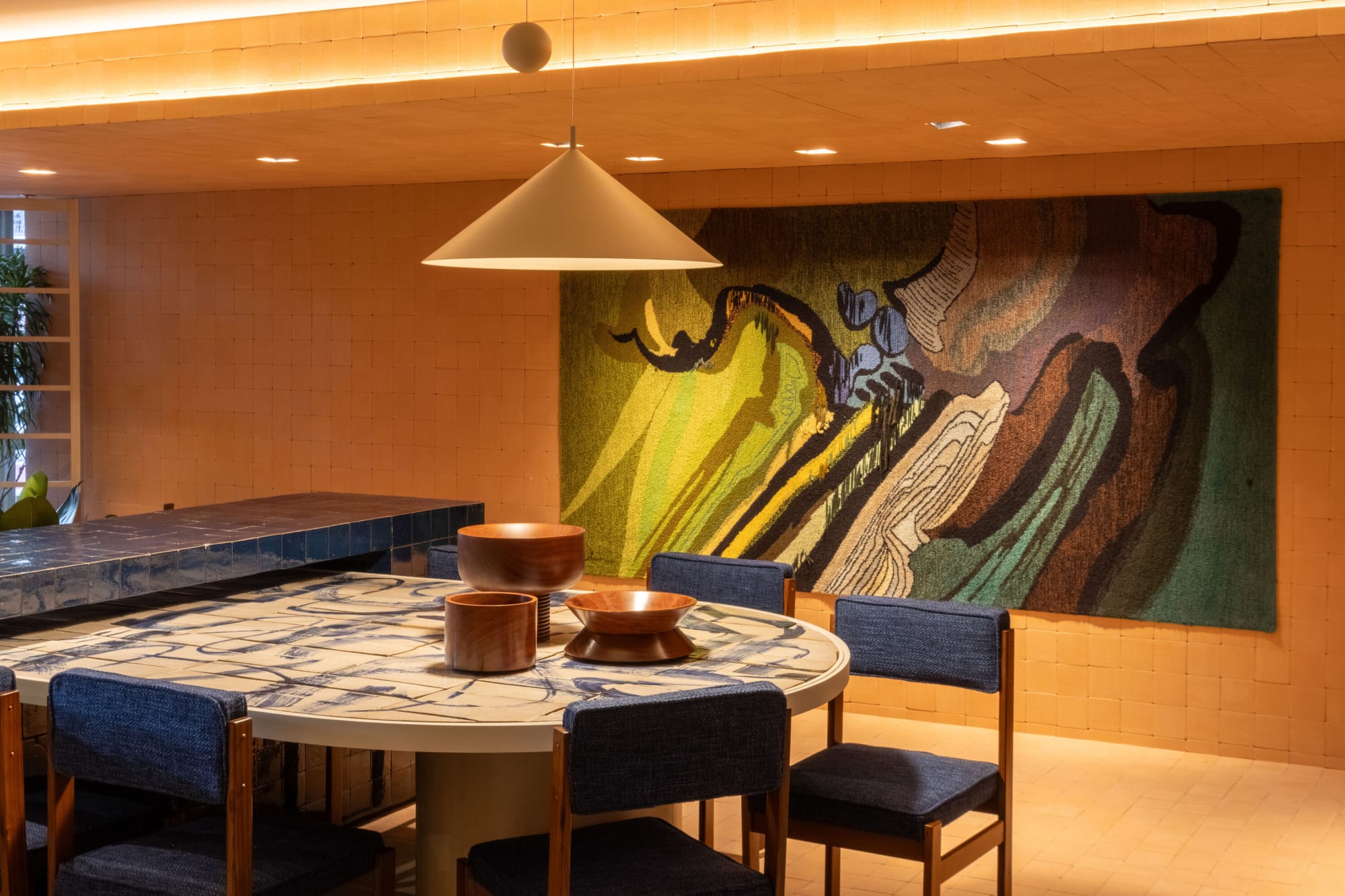 Round dining table with blue upholstered chairs under a cone lamp and abstract mural