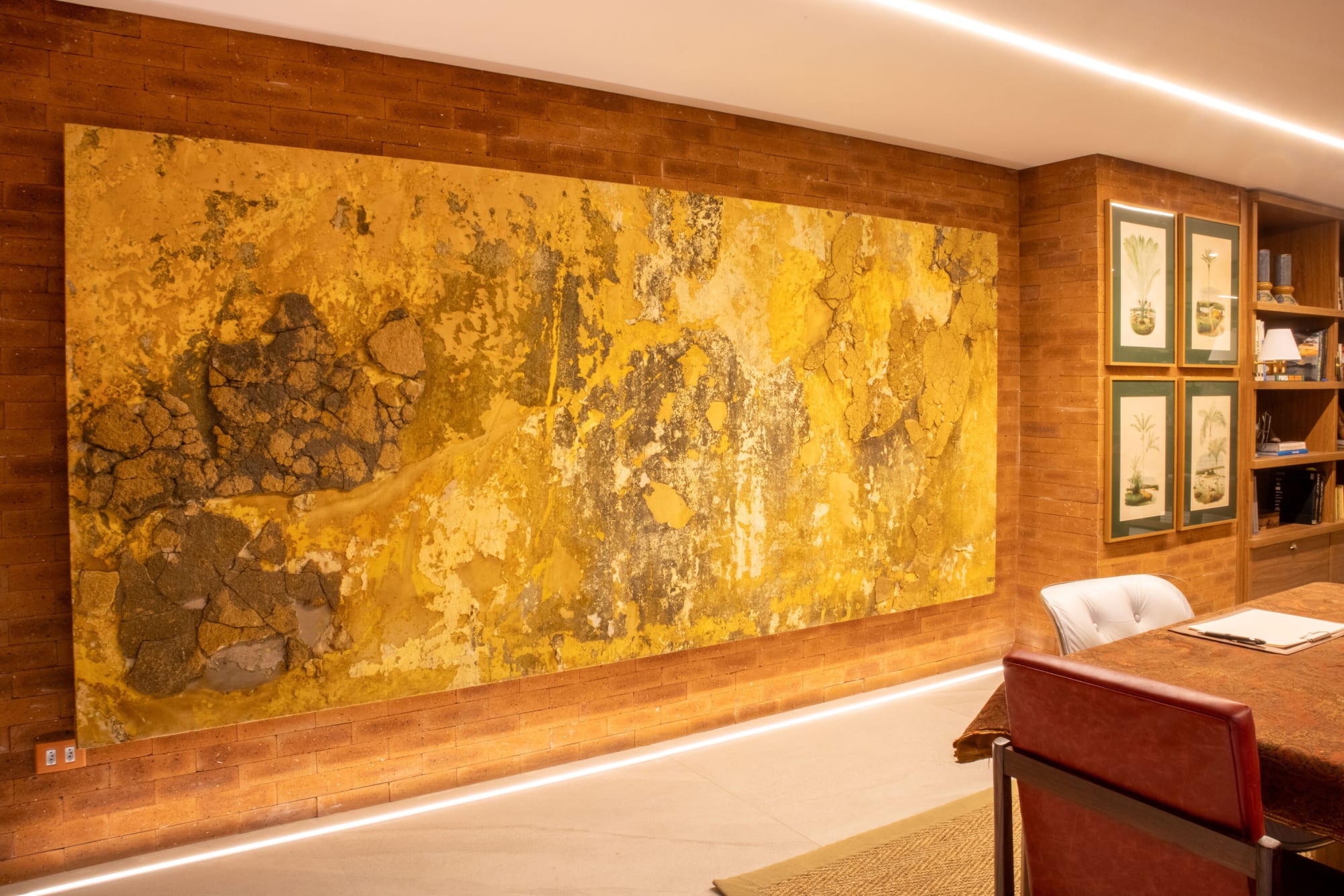 Large gold textured painting across a clay brick wall beside framed botanical prints