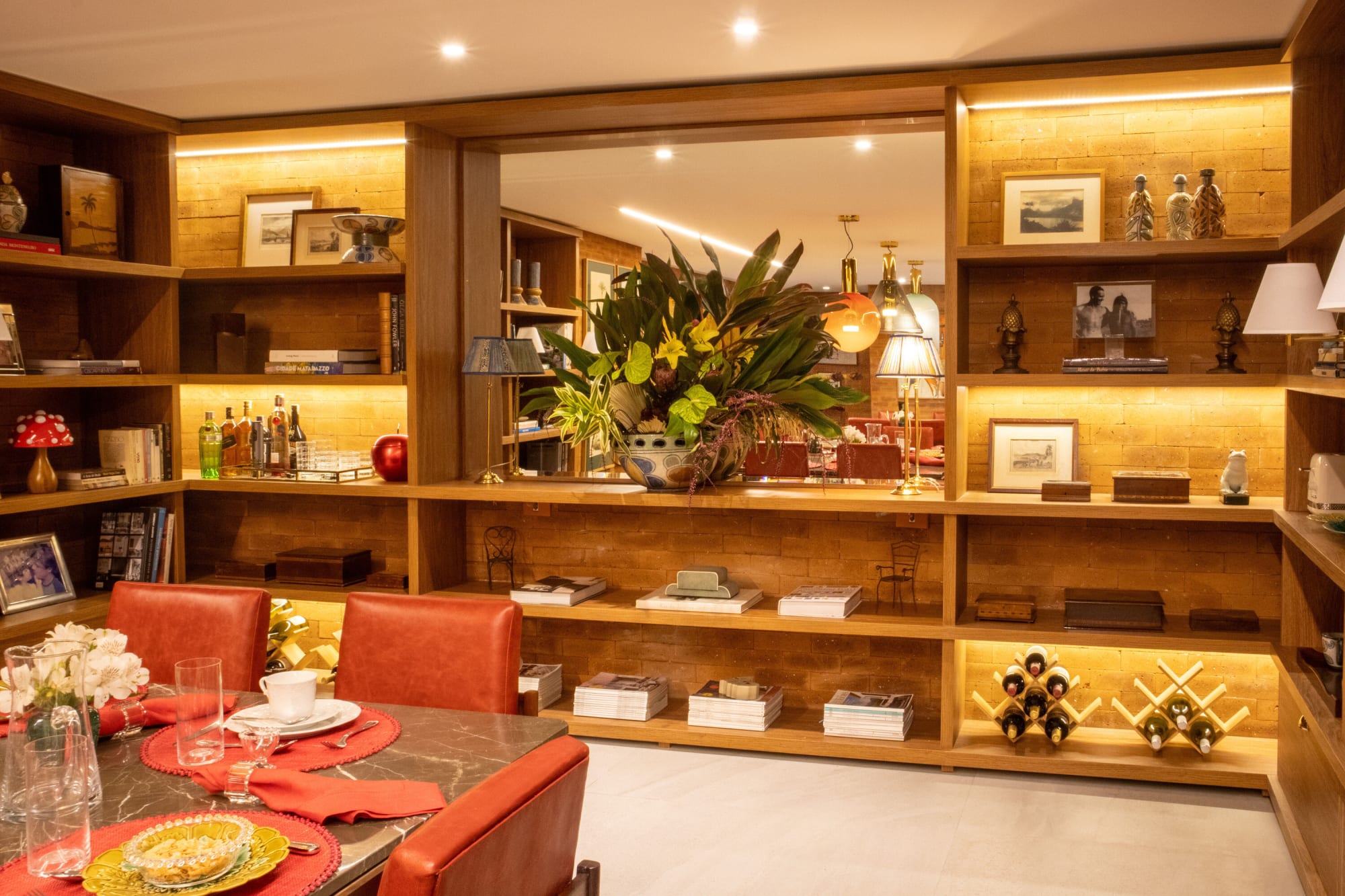 Red chairs at a laid table facing oak shelving and a mirror reflecting tropical flowers
