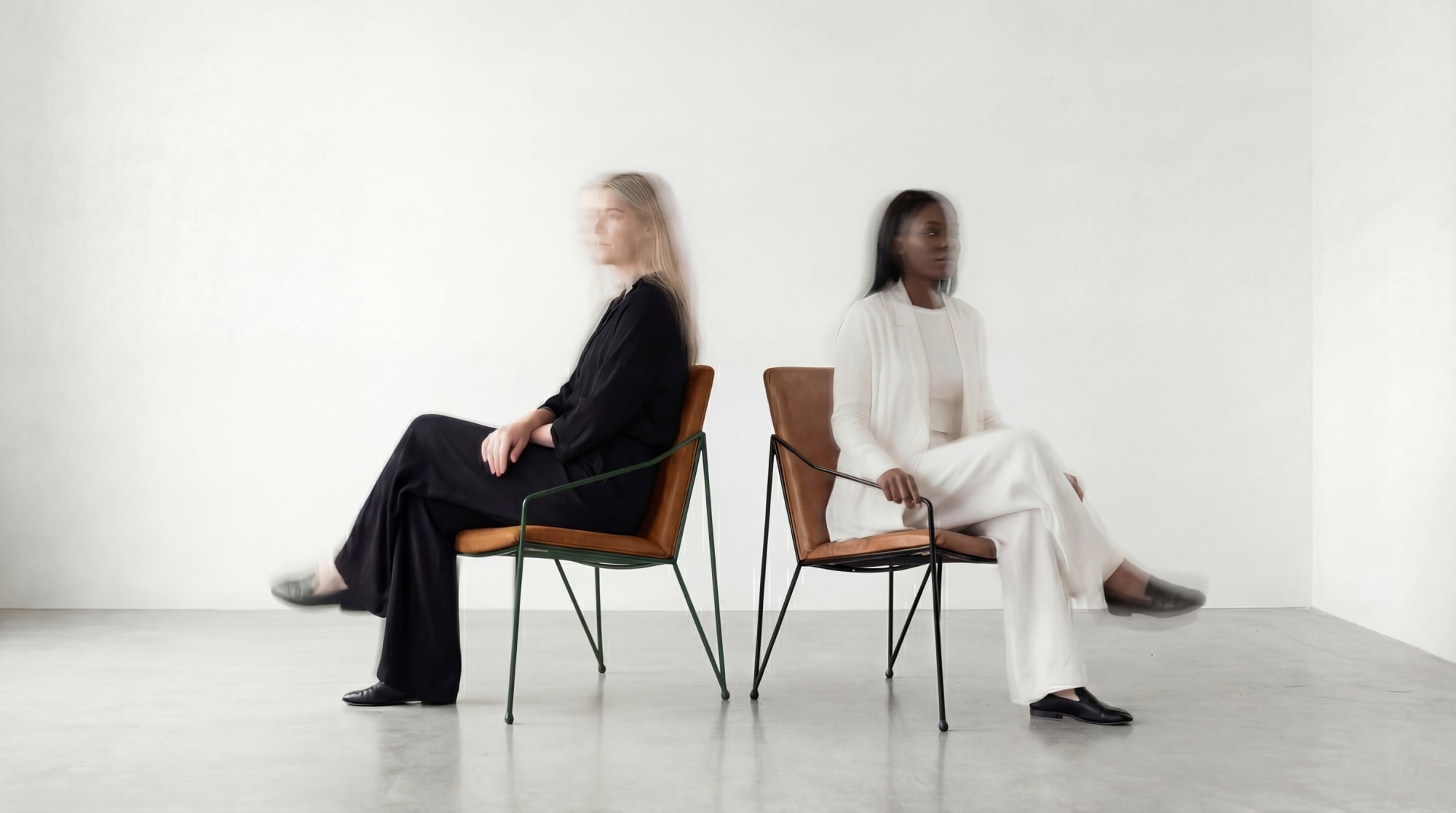 Two models in black and white suits seated on tan leather Cremme chairs in a white studio