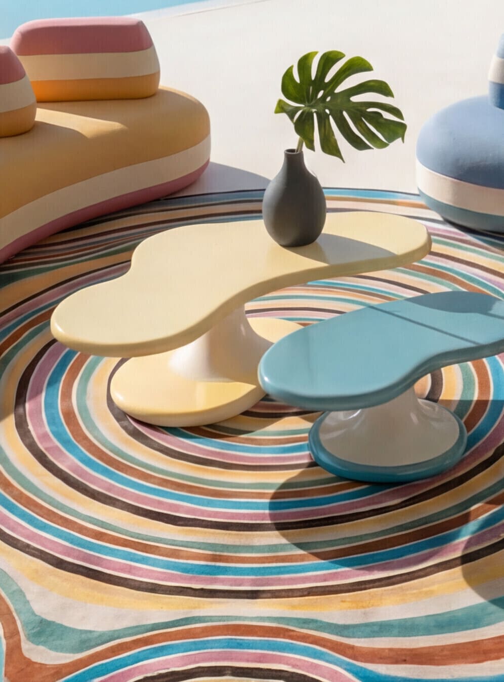 Pastel yellow and blue curved coffee tables on a striped swirl rug with a monstera vase