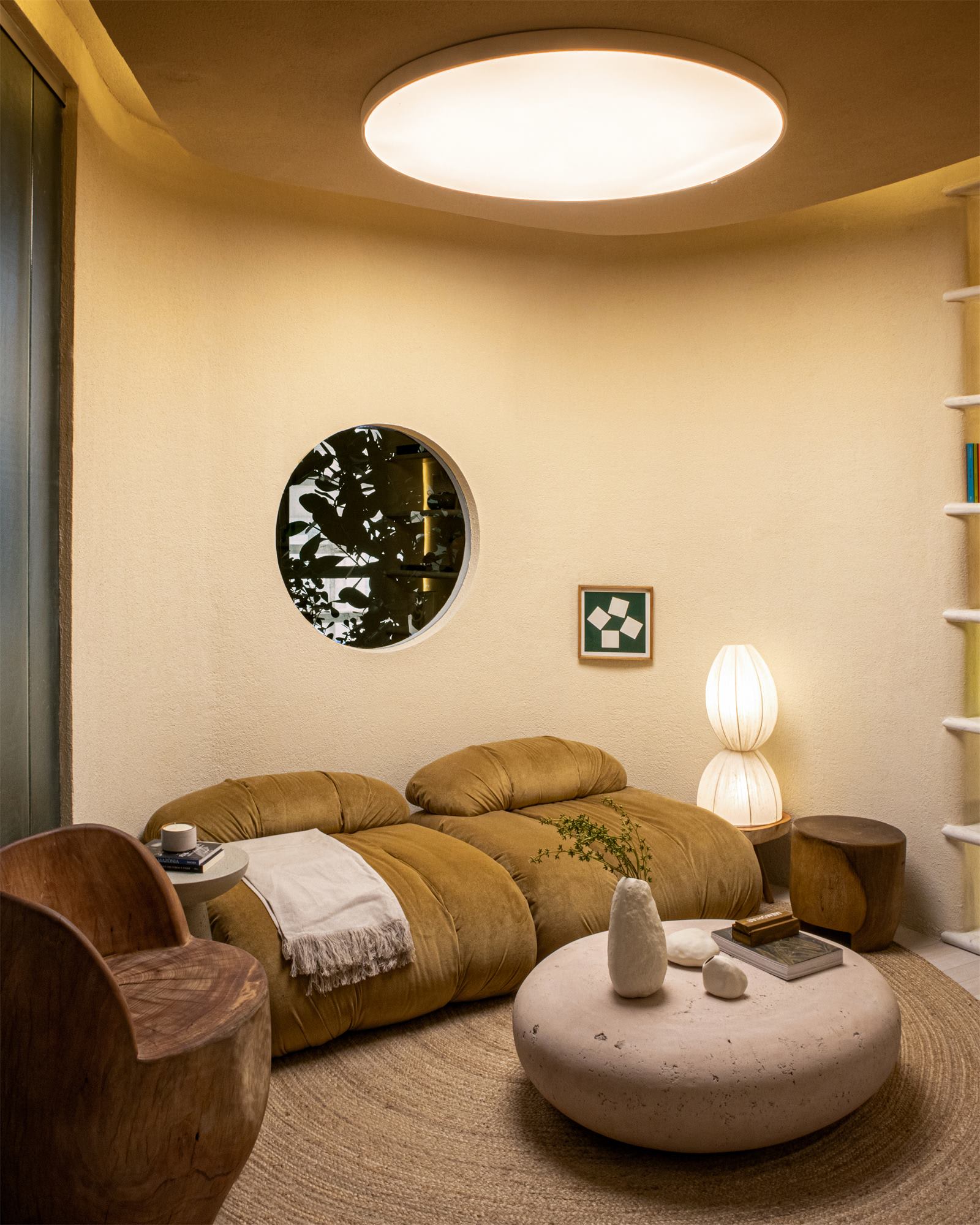 Round reading room with an ochre velvet sofa, a stone coffee table and a porthole window onto green leaves