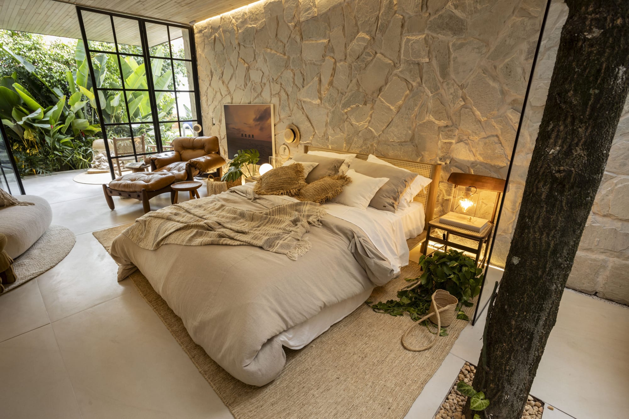 Cama con manta tejida contra un muro de piedra blanca junto a un tronco de árbol