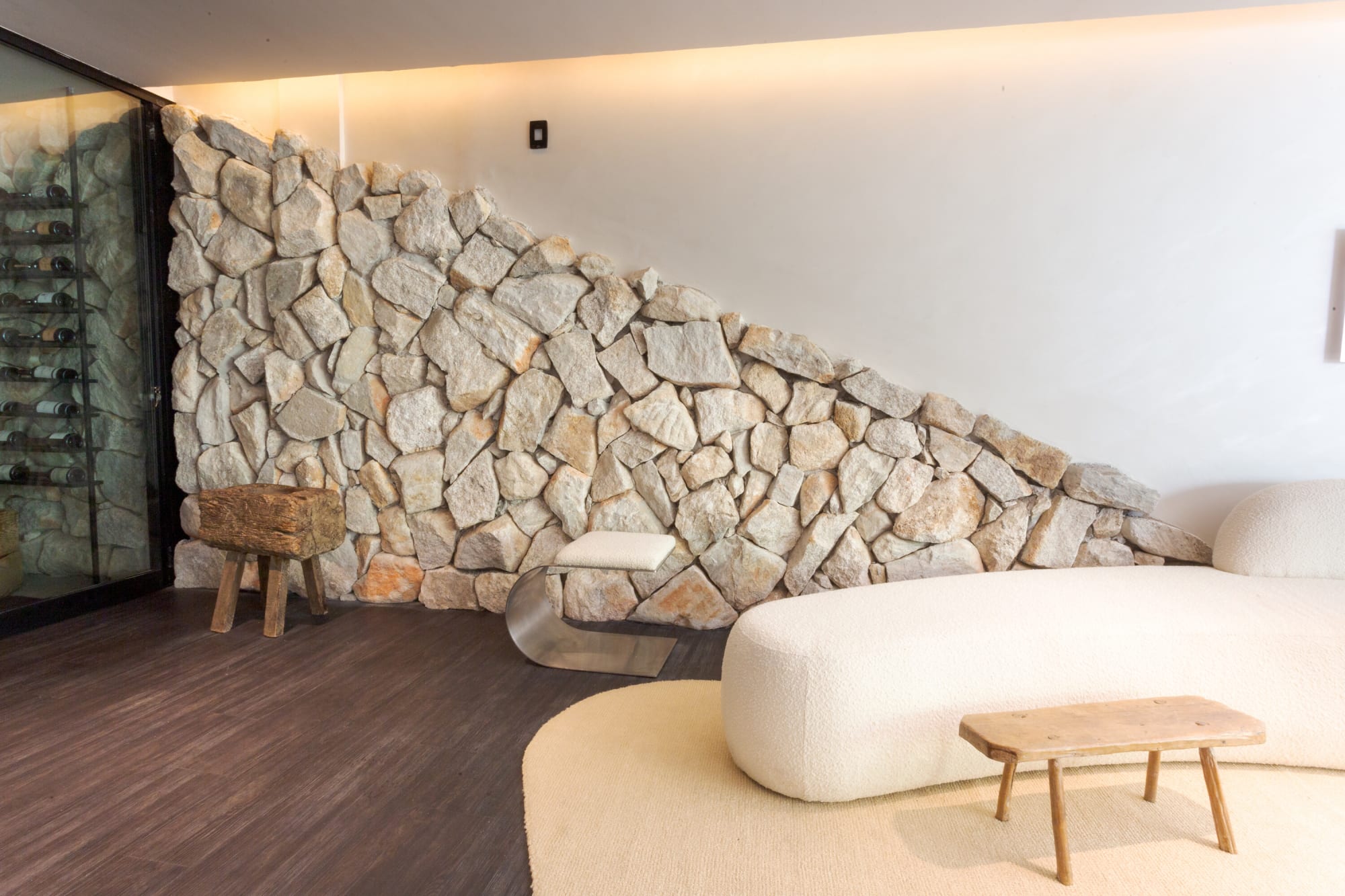 Rough stone wall rising over a white sofa and a small timber bench on a dark floor