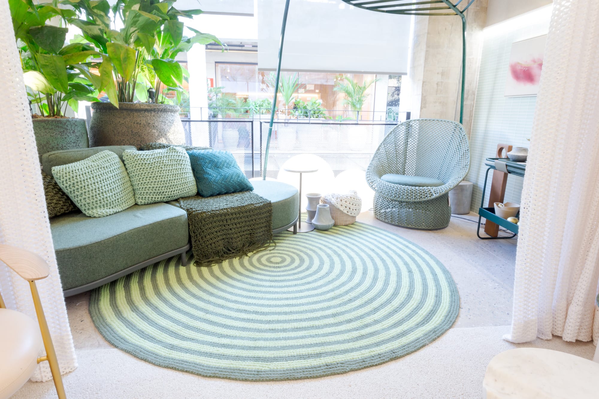 Sage sofa and woven egg chair on a green striped rug before glass walls and planting