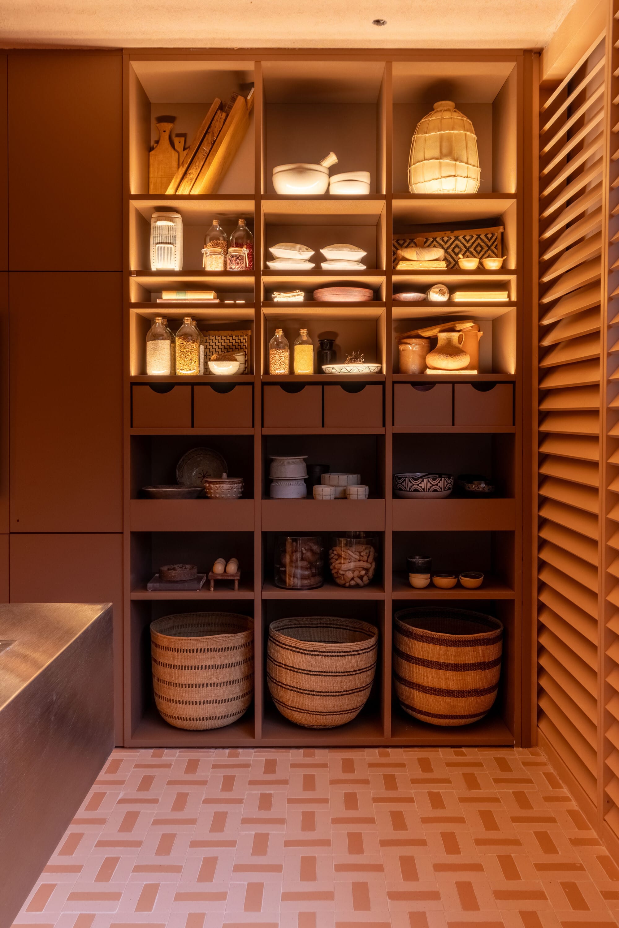 Lit kitchen shelving with ceramics, jars and woven baskets beside timber shutters