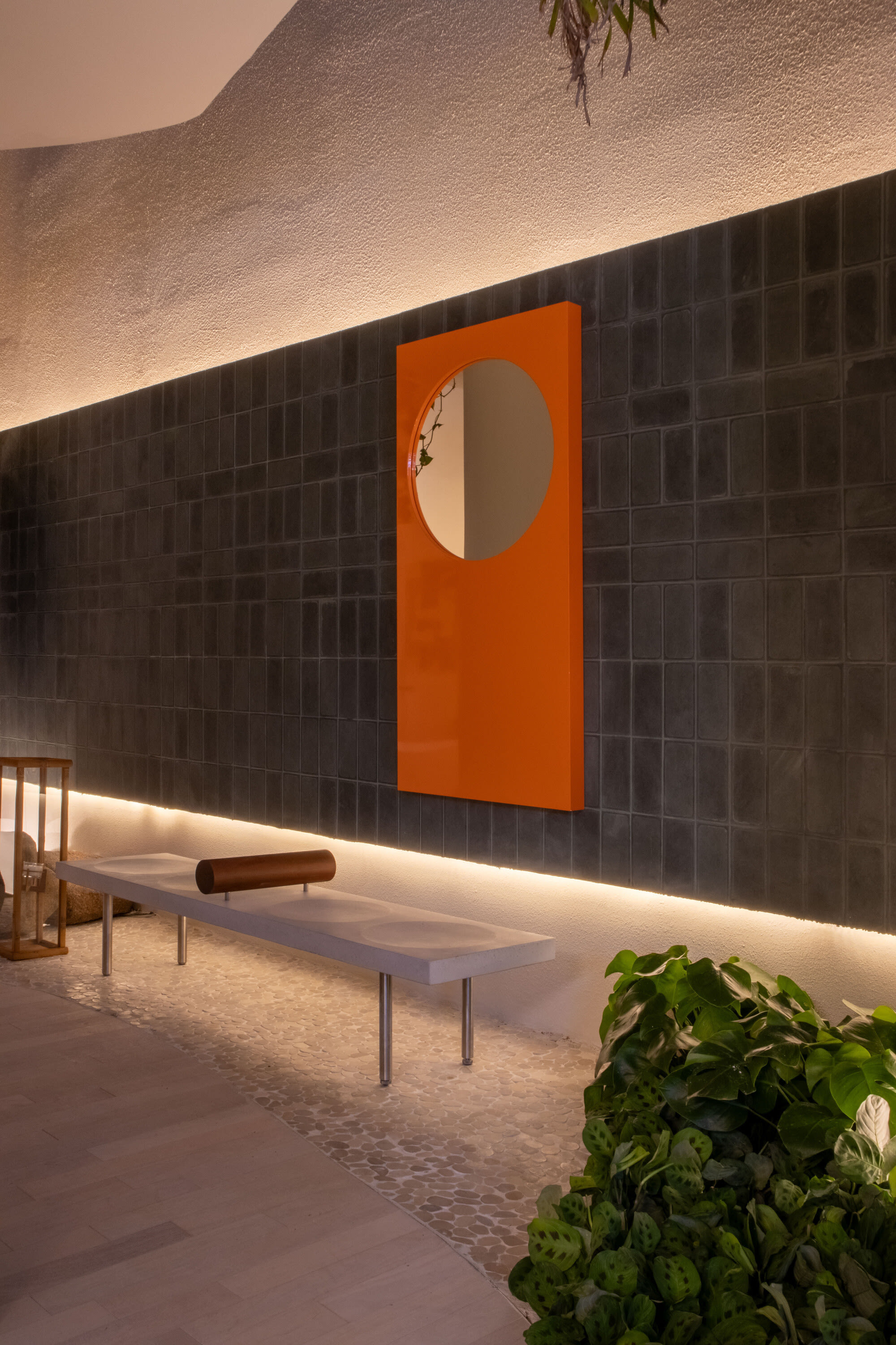 Orange panel with round mirror on a dark tiled wall above a white bench and pebbles
