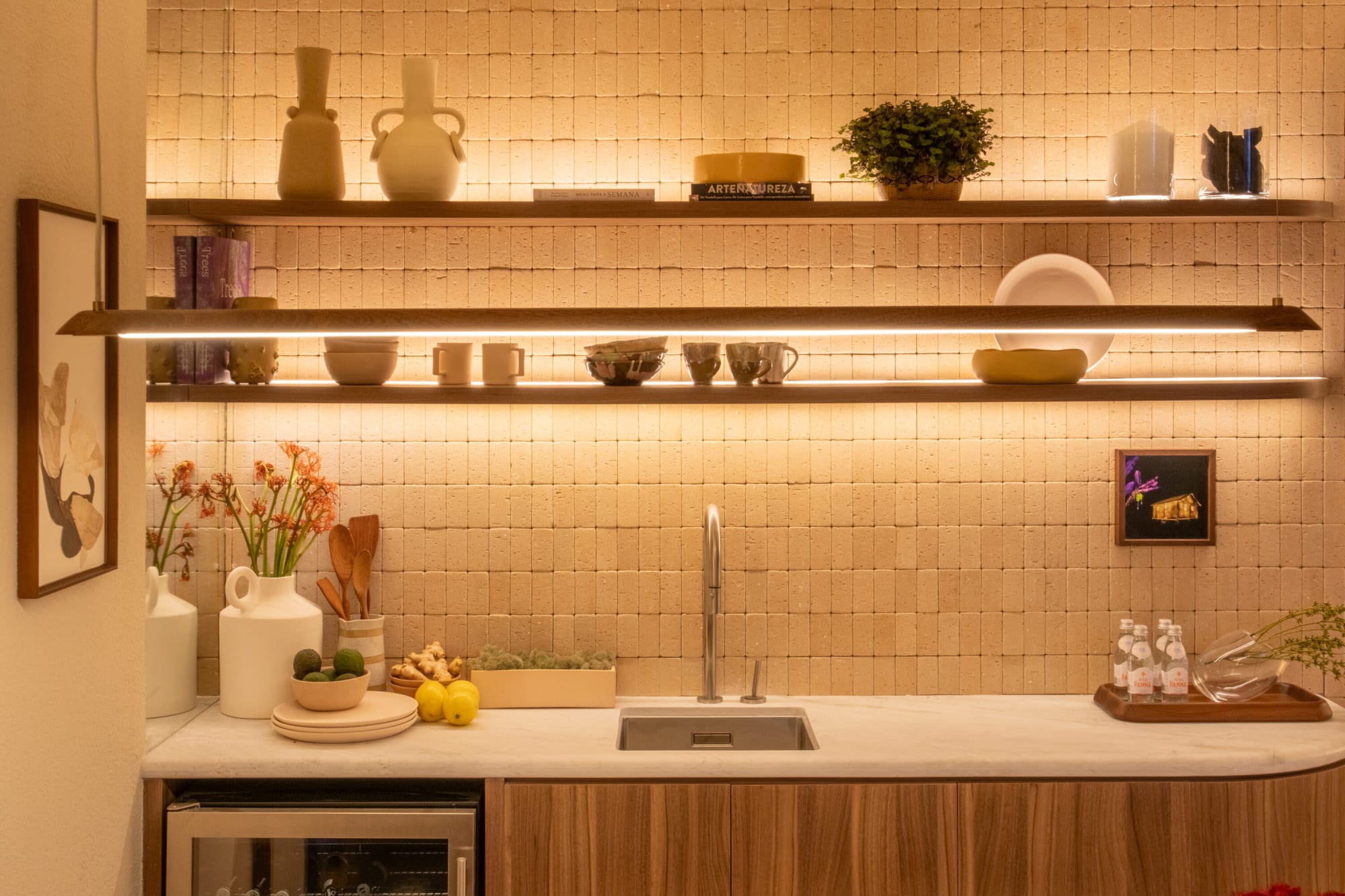 Lit open shelves with ceramics and plants above a kitchen sink and stone counter