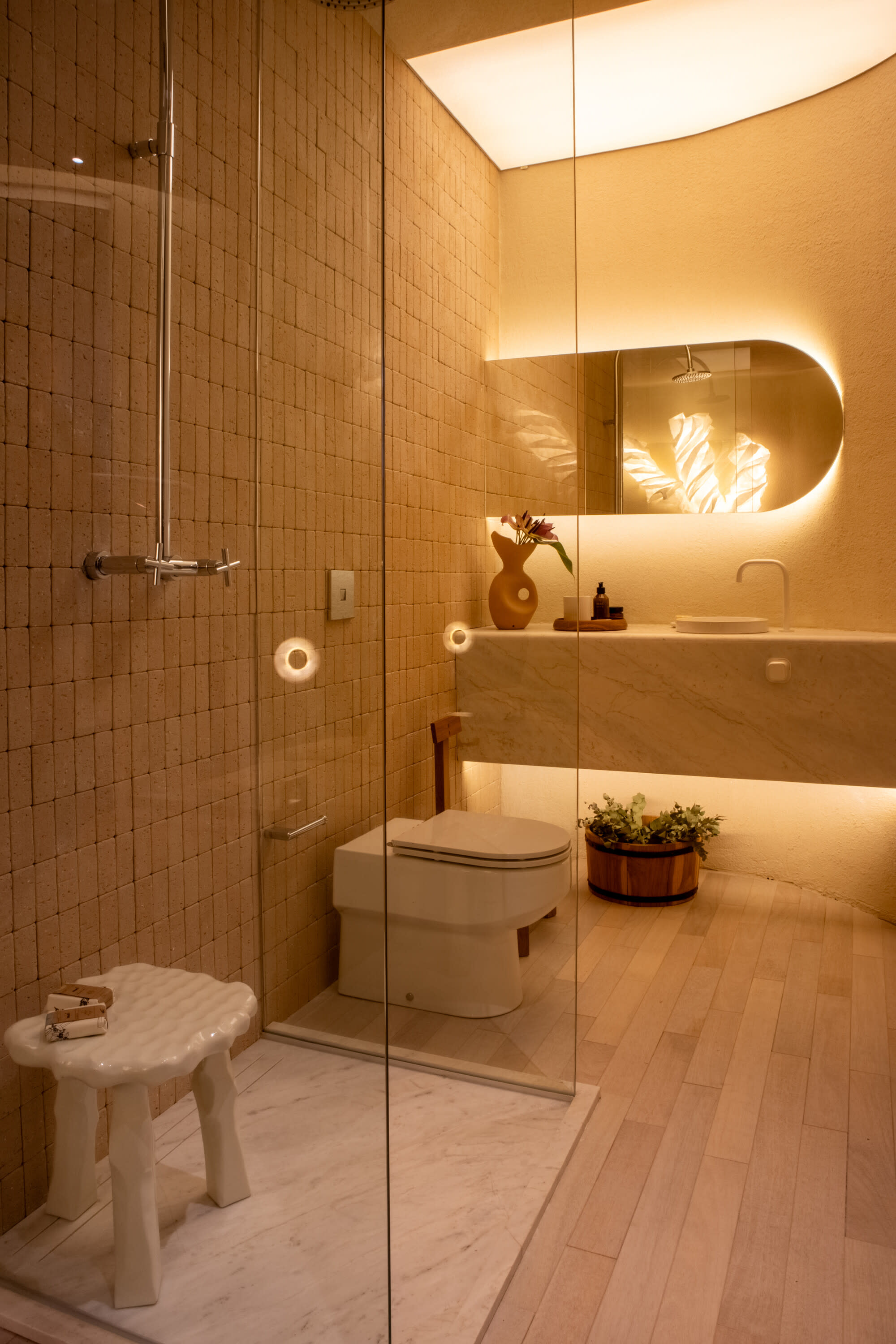 Glass shower with tiled walls, white stool, toilet and a backlit oval mirror