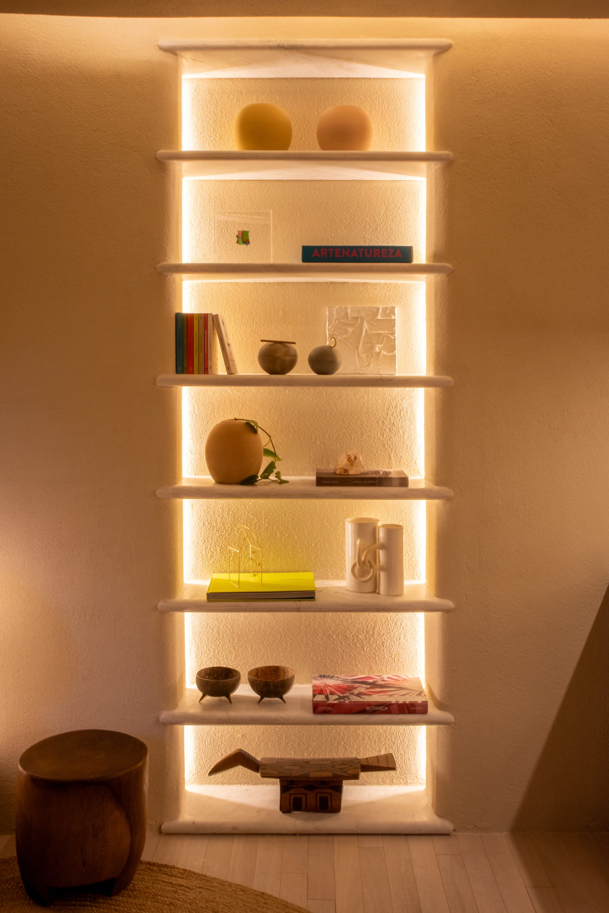 Tall lit niche of shelves holding ceramics, books and a carved timber stool