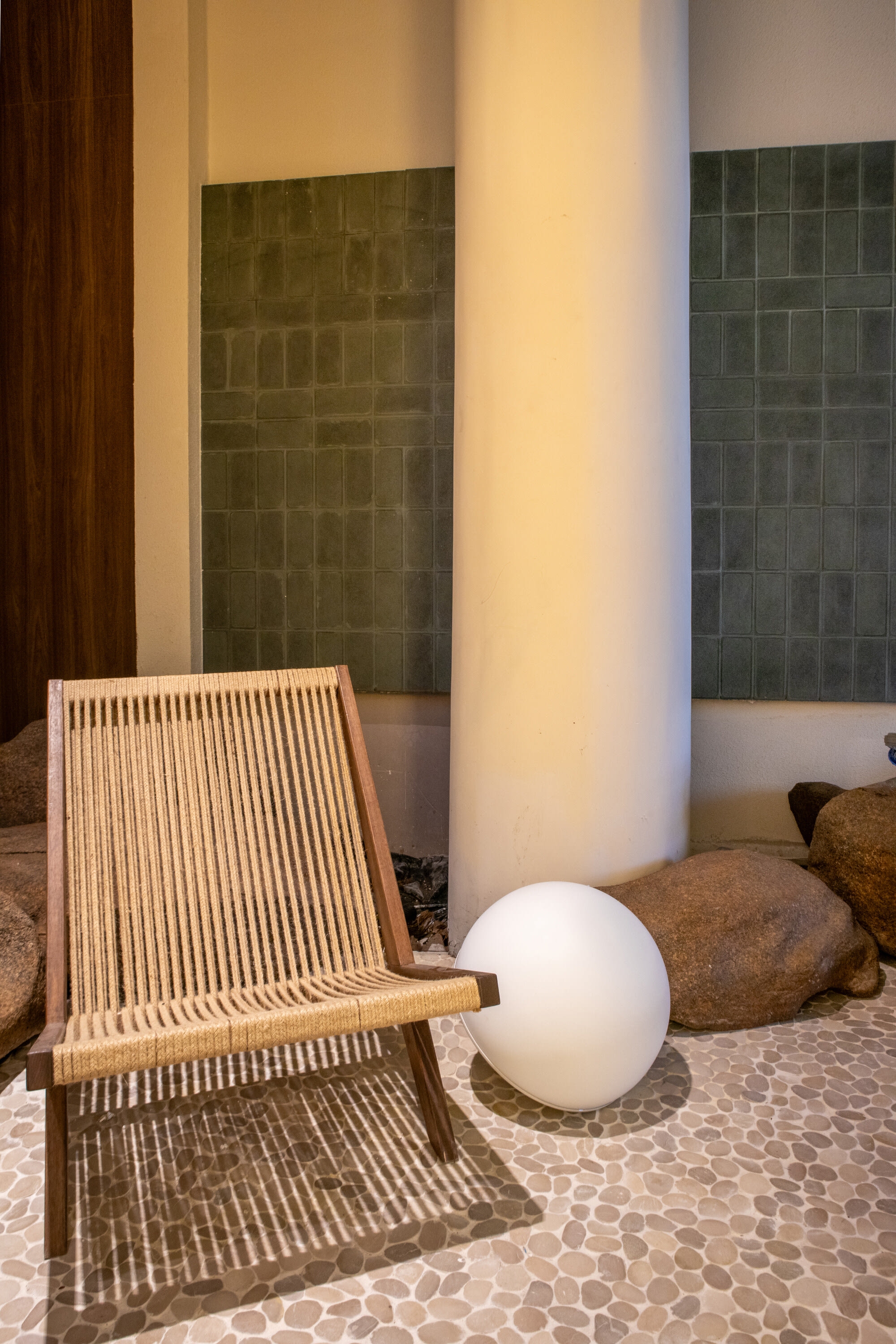 Rope sling chair and white globe lamp on a pebble floor beside a cream column