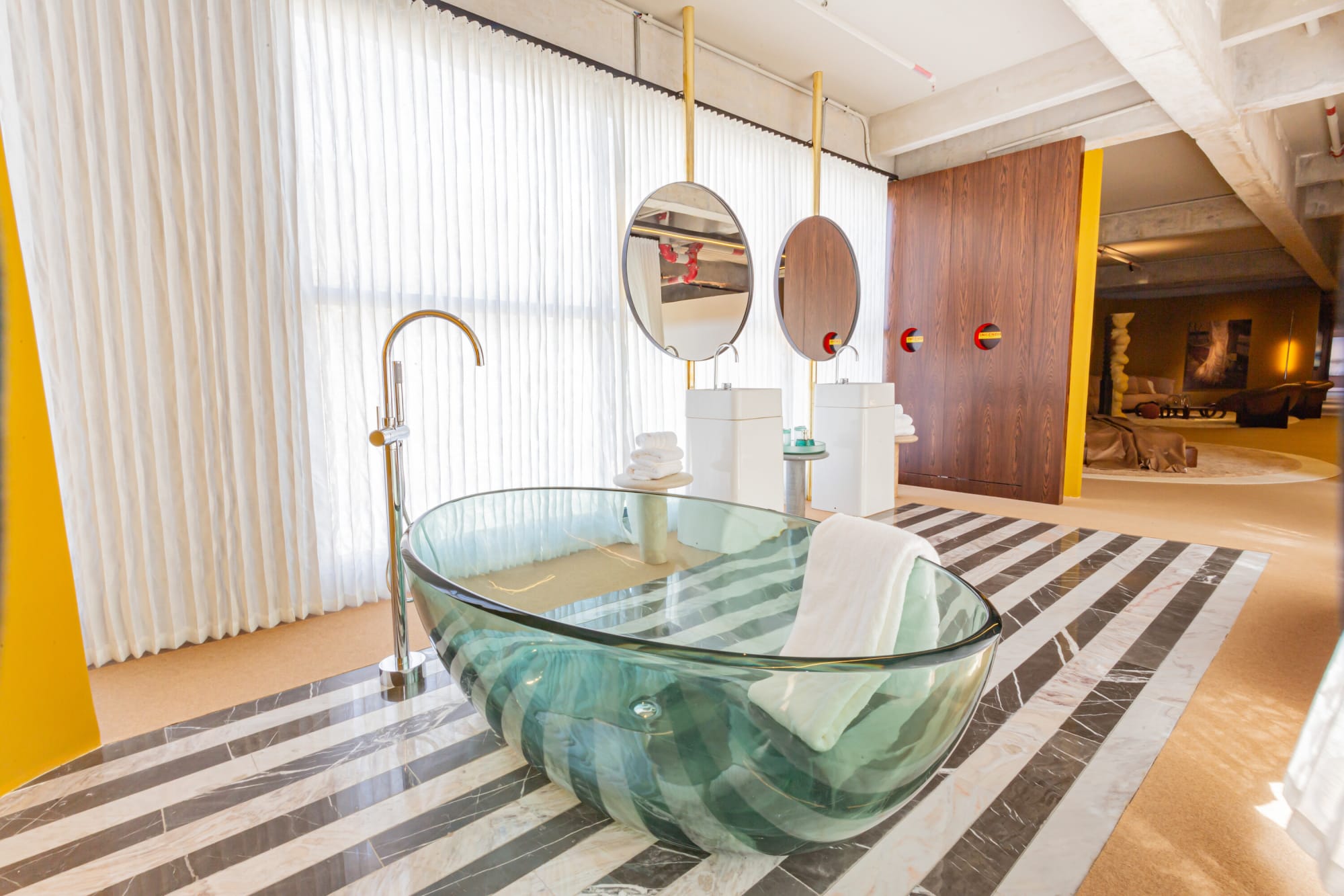 Glass bathtub with floor tap on striped marble before sheer curtains and round mirrors