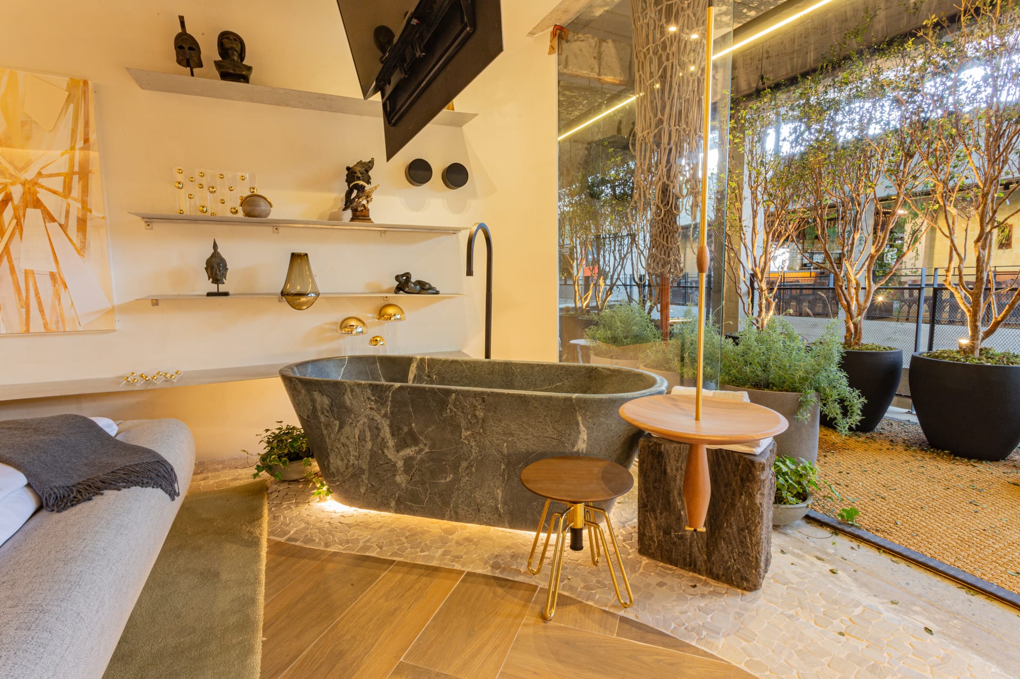 Dark stone bathtub with brass tap and a timber stool beside potted trees and shelves