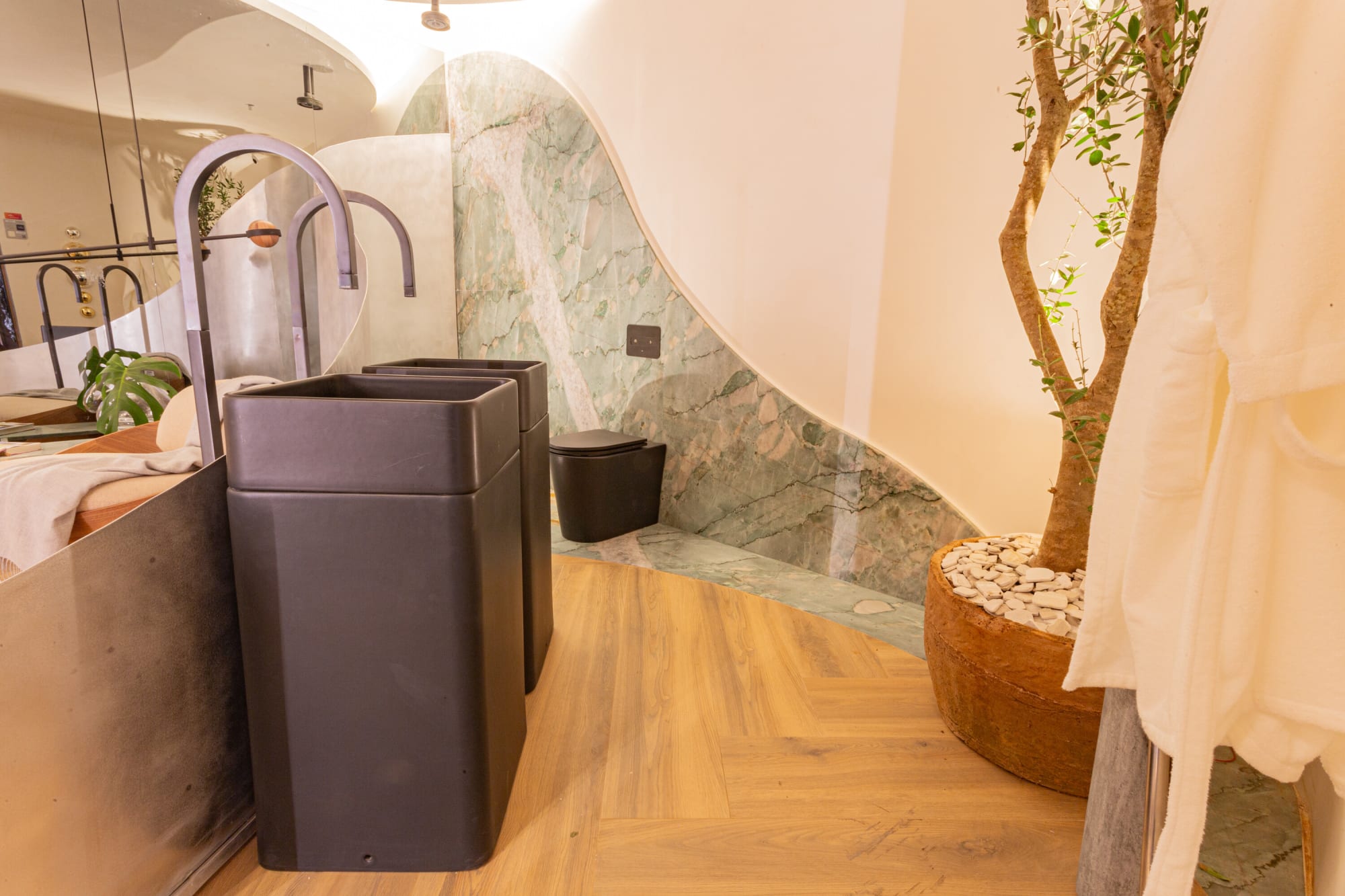 Two dark pedestal basins with tall black taps beside green marble and a potted tree