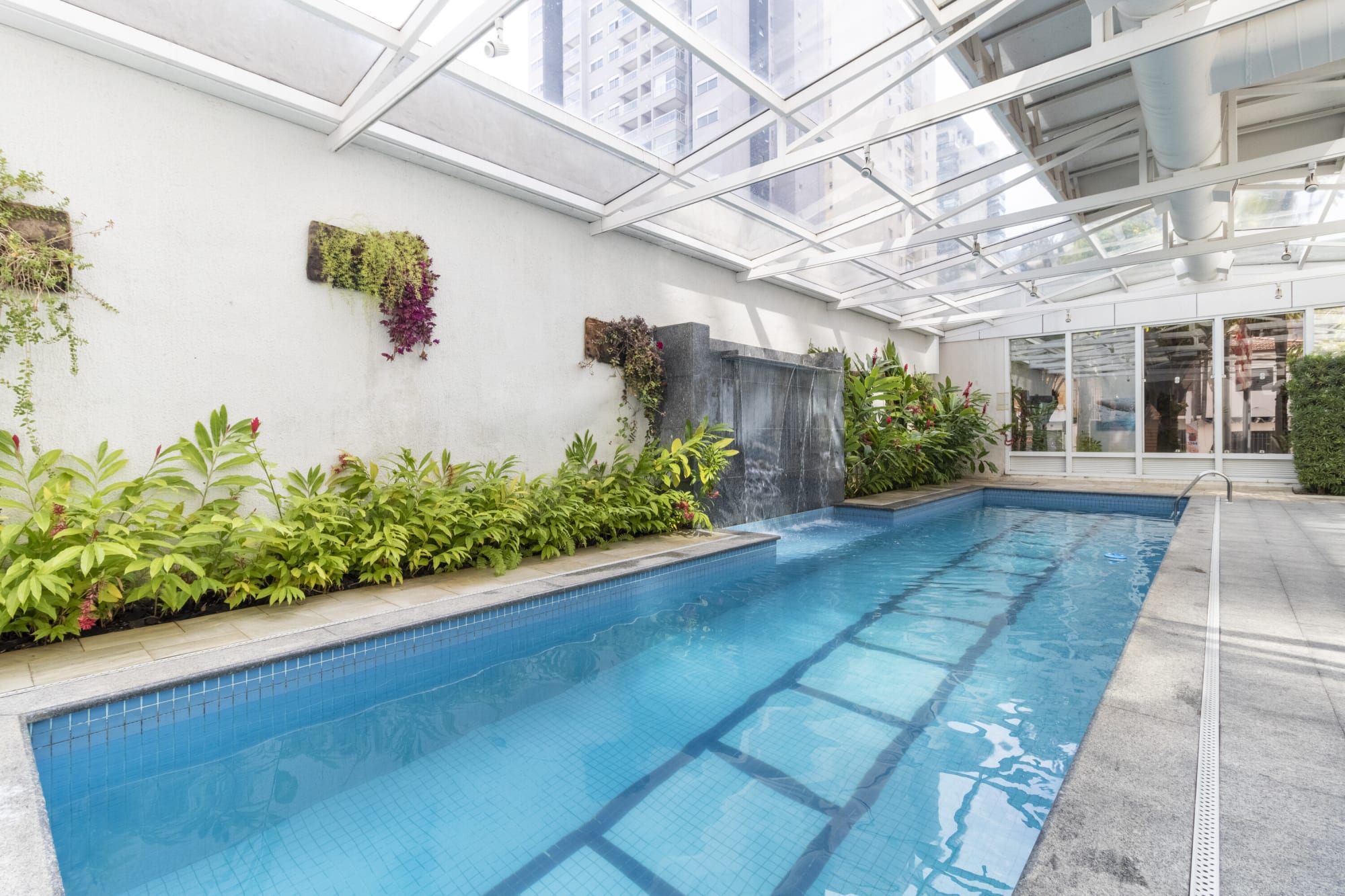 Long blue pool beside a wall waterfall and planted bed under glass