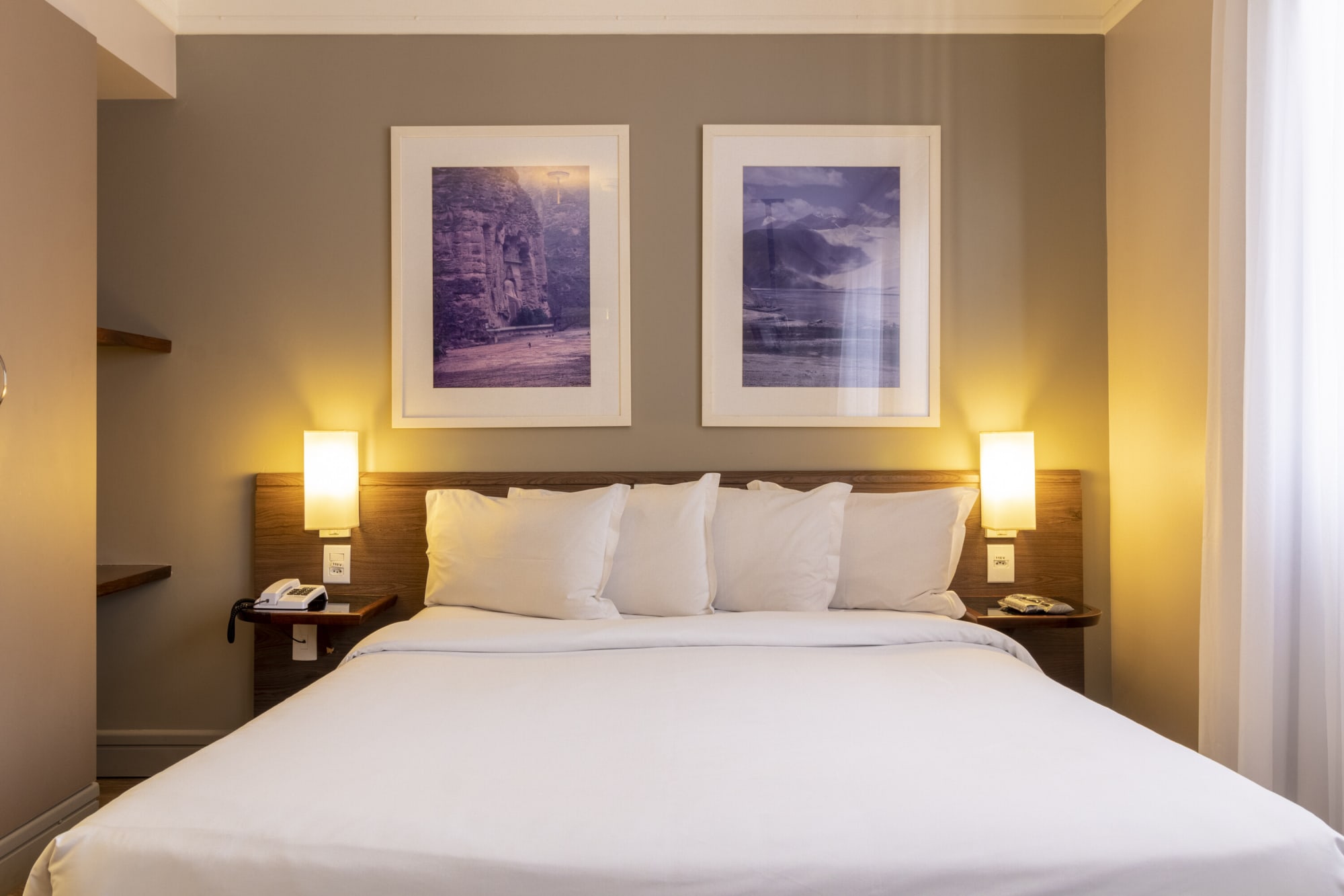 Hotel bed with white pillows under two blue framed prints and wall lamps